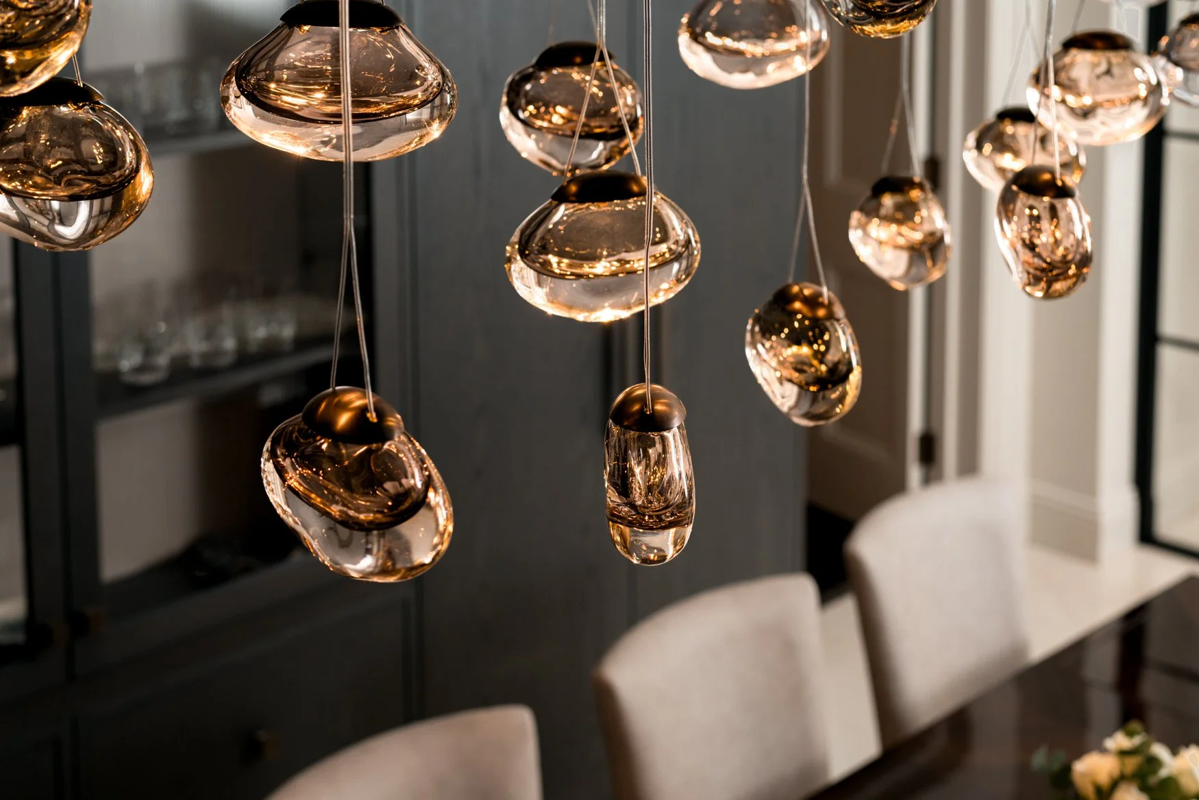 A close-up interior detail photographed in London, showing warm amber pendant lights above a dining area with dark joinery and soft neutral seating in the background. The image captures the mood, reflection, material finish and atmosphere created by 