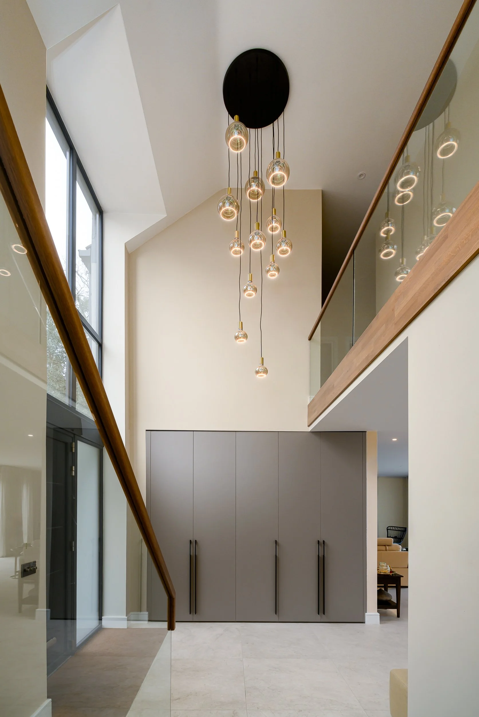 Interior architecture photograph of a double-height residential space with full-height glazing, glass balustrade, staircase, fitted storage and suspended feature lighting in a luxury home in London.