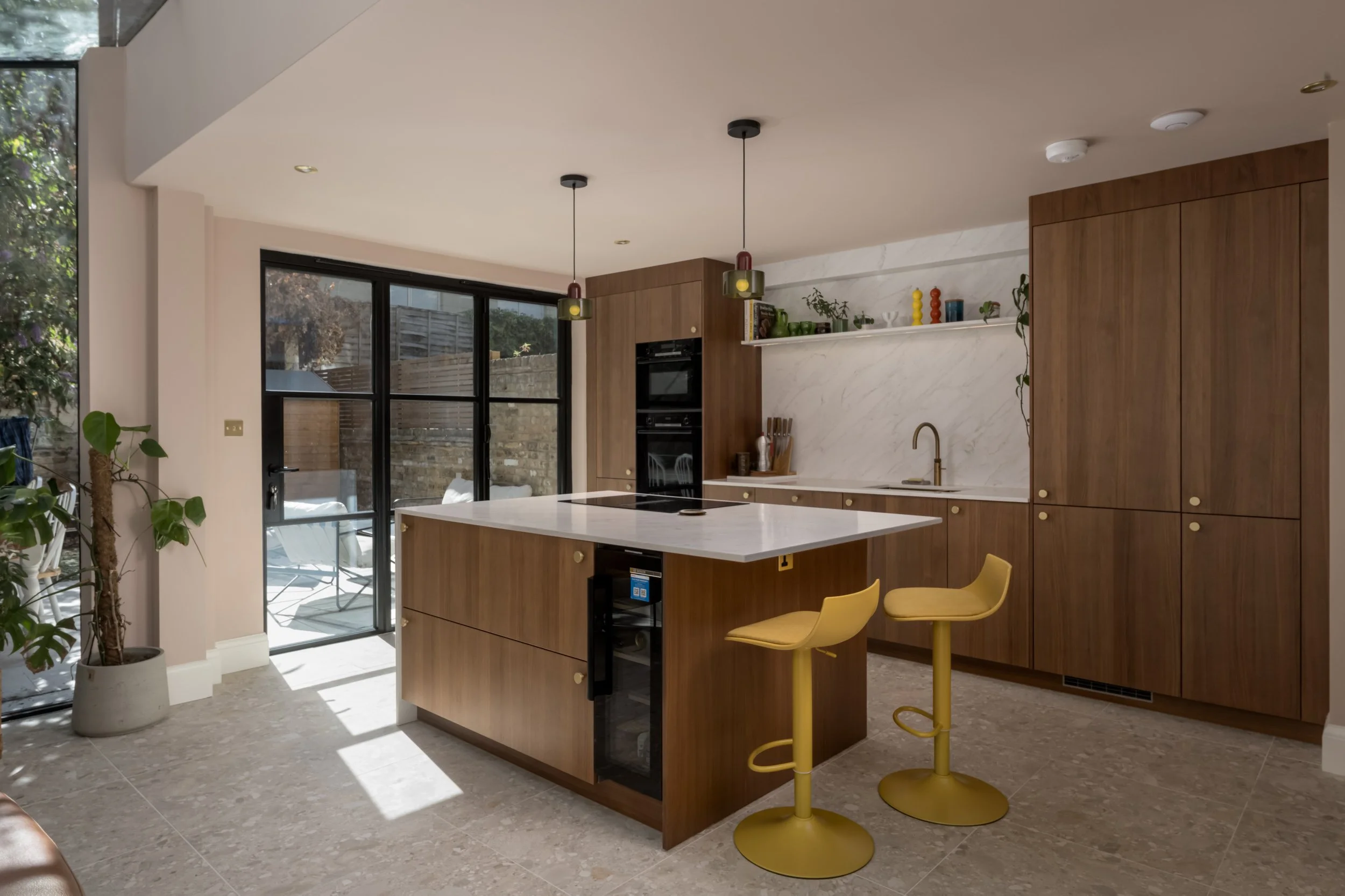 Modern kitchen with wooden cabinetry, white marble backsplash, and a kitchen island with a white countertop. Two yellow bar stools are at the island. Large glass doors lead to an outdoor patio with chairs. Potted plants are inside near the door, and 