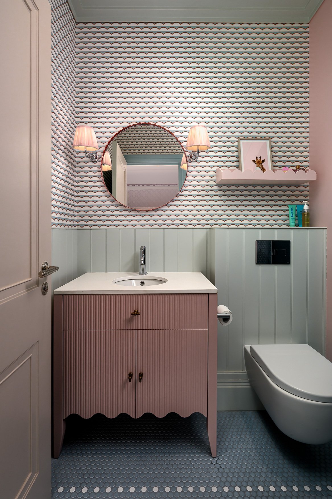 Children’s bathroom vanity with pink cabinet, round mirror, wall lights and patterned wallpaper.