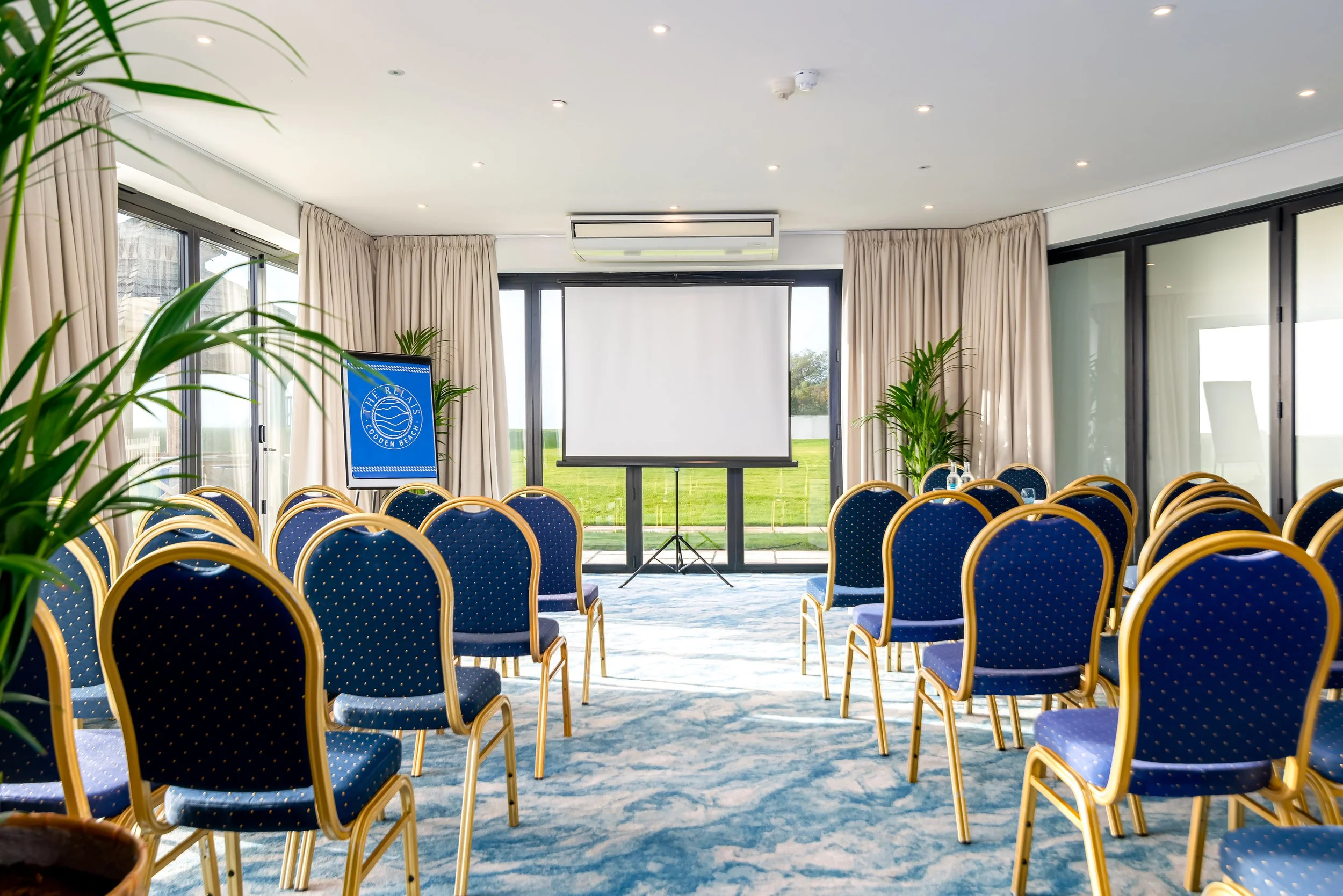 Conference room with rows of blue chairs with gold trim facing a projection screen, decorated with potted plants and large windows with beige curtains.
