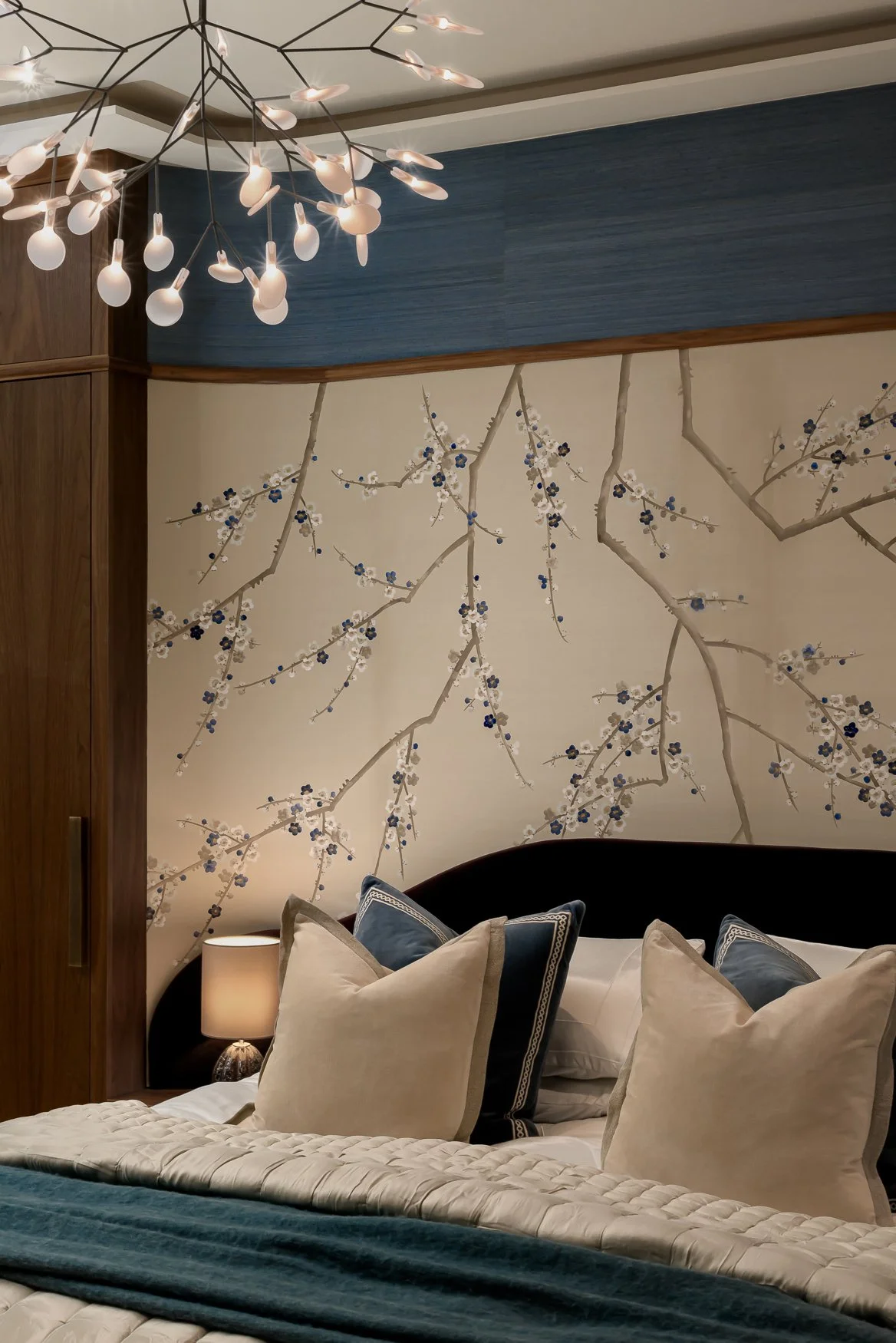A bedroom with beige curtains, a black headboard, multiple pillows, a floral wallpaper with white and dark blue blossoms and branches, a wooden cabinet, a bedside lamp, and a modern starburst chandelier with small white lights.