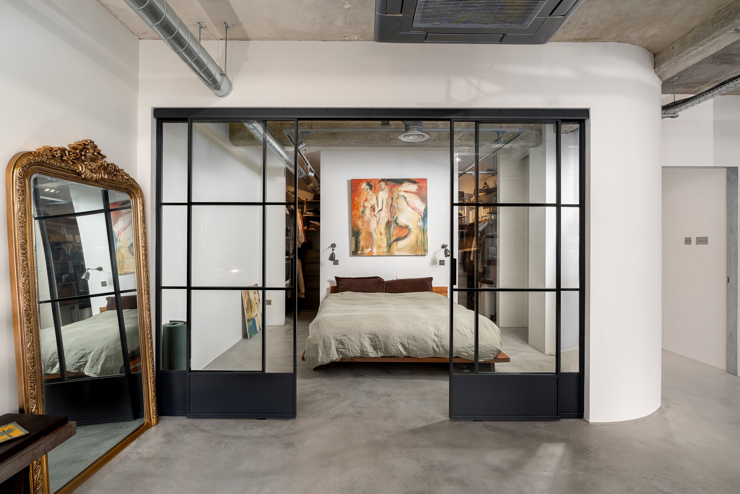 Interior architecture photograph of an industrial-style loft bedroom with black framed glass partition doors, exposed concrete ceiling, visible ducting and curved wall detail.