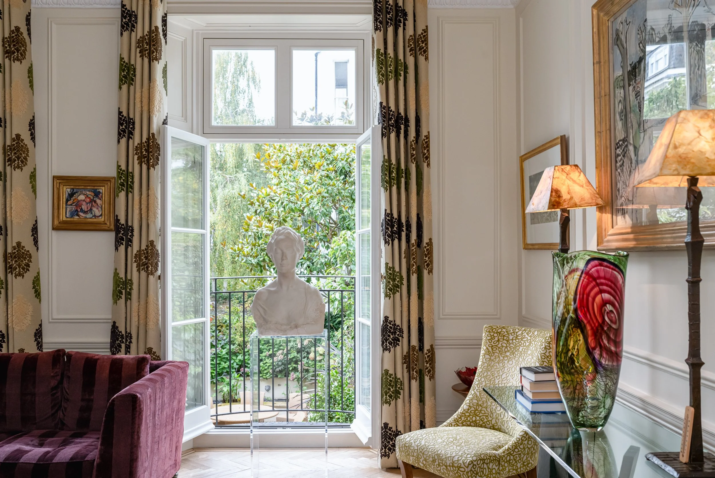 An elegant London townhouse interior with French windows opened towards a leafy garden view. The image highlights the tall glazing, natural light, classic proportions and how the windows frame the connection between the interior and outside space.