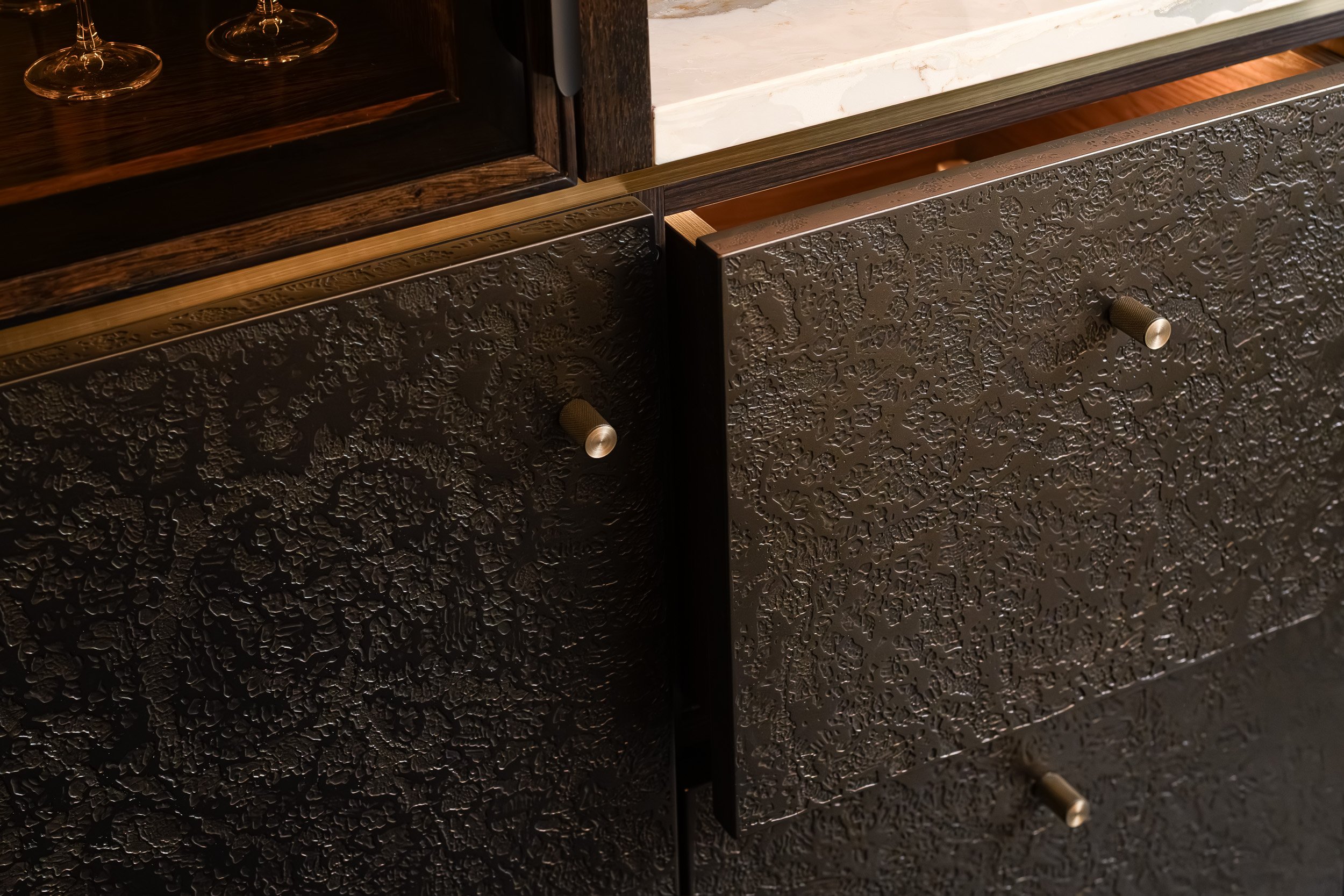 Close-up of a black textured cabinet with metallic drawer pulls, part of a furniture piece with a white marble top and glasses with wine on top.