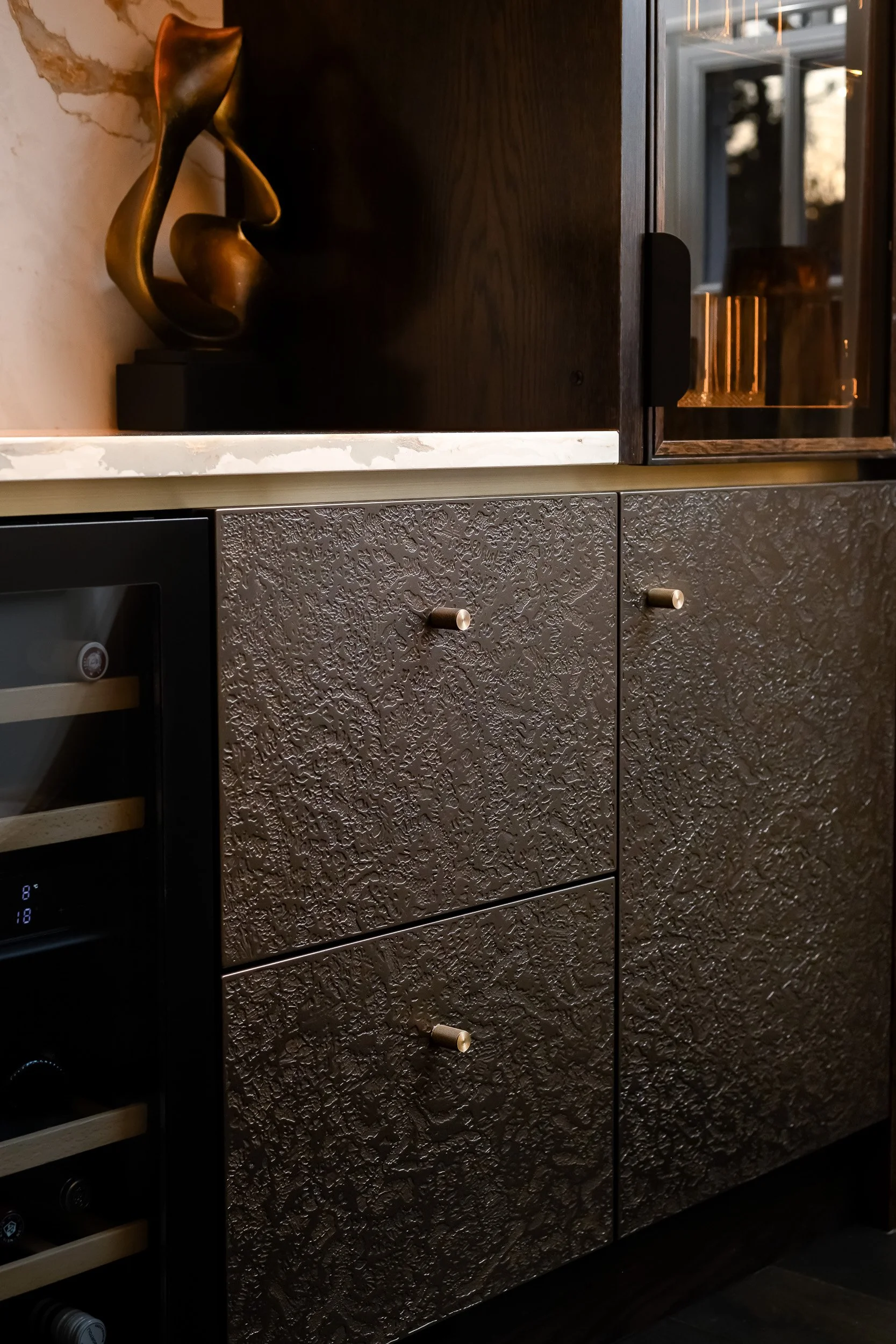 A modern kitchen cabinet with a textured metallic finish and small round knobs, situated below a dark wooden countertop with a decorative bronze sculpture on top, and a wine cooler on the left side.