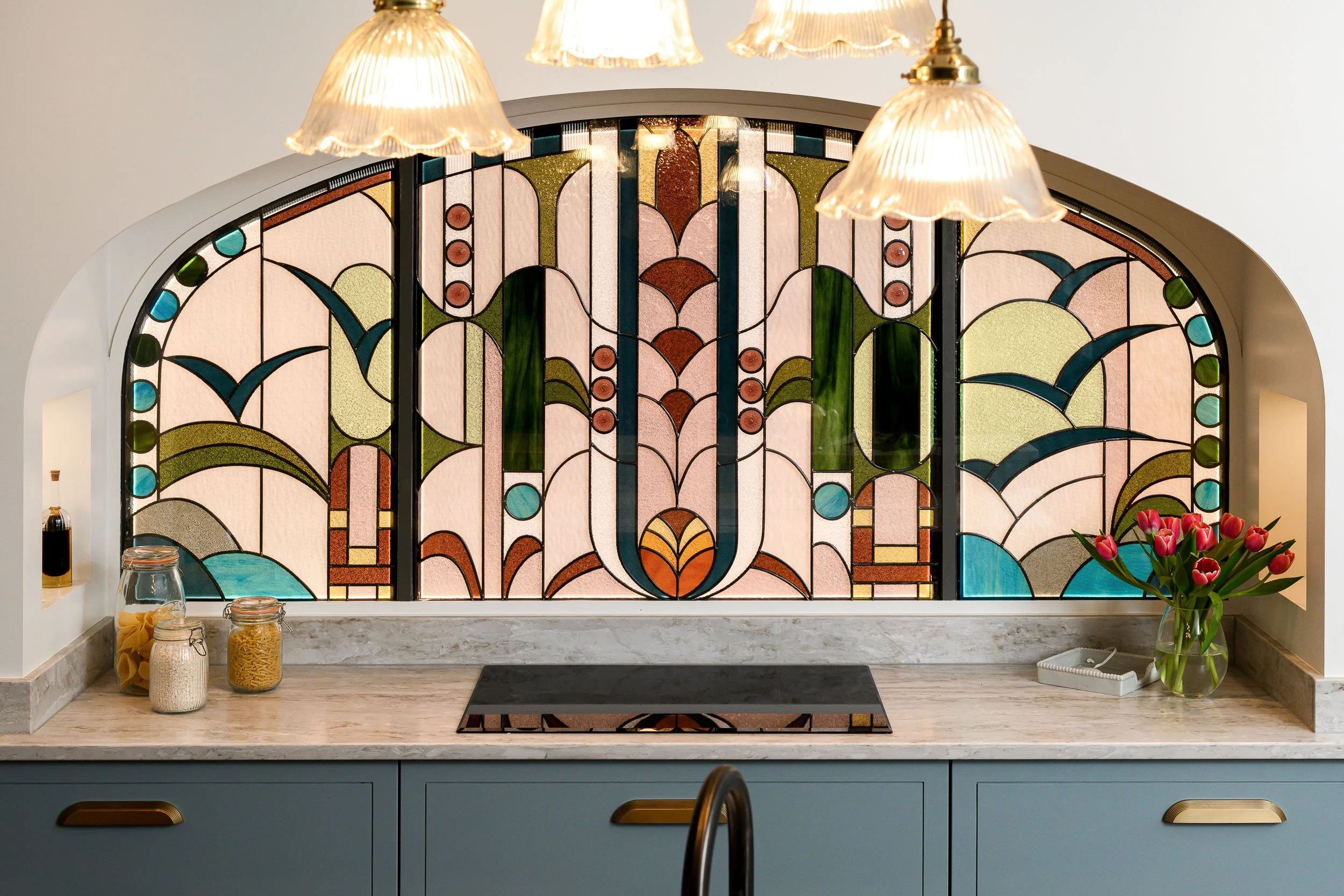 Bespoke kitchen with a colourful decorative splashback framed by an arched opening and pendant lights.
