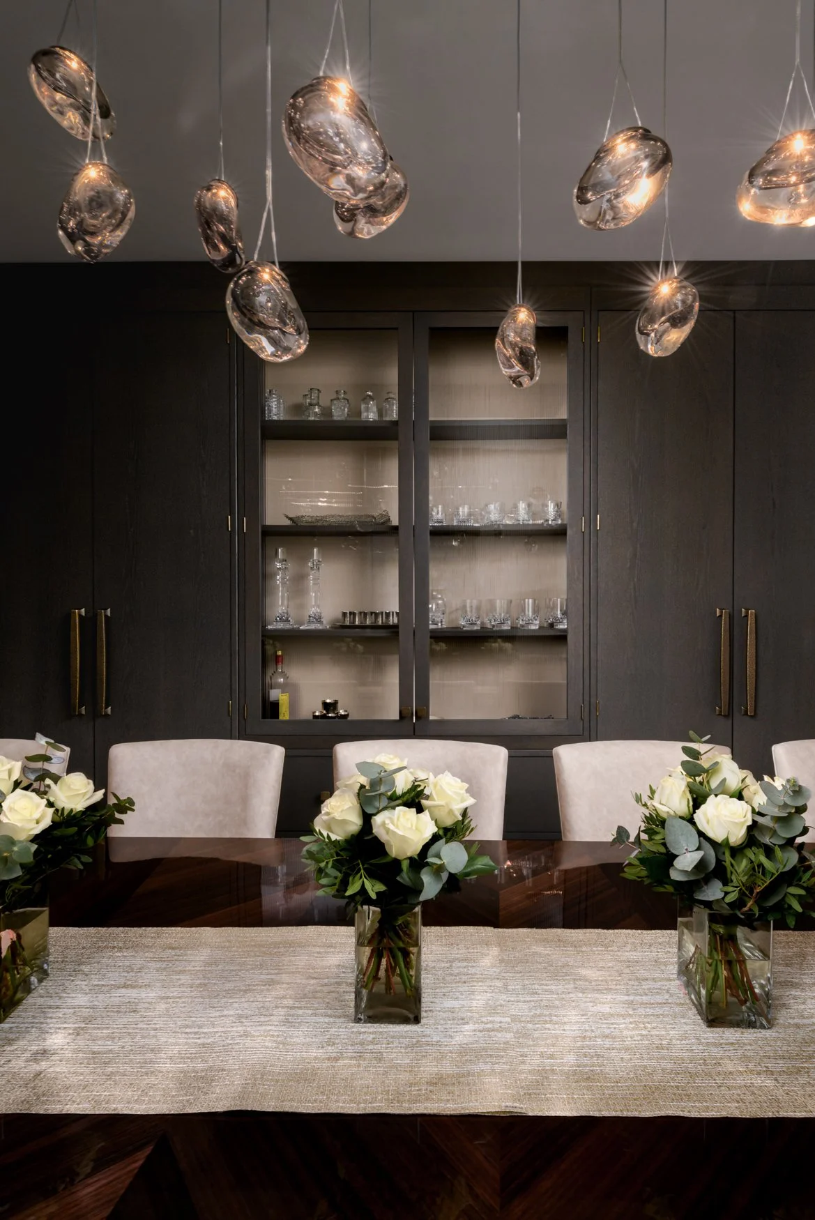 A dining room with a dark wooden cabinet displaying glassware, set against a dark wall, and decorated with three vases filled with white roses and greenery on a wooden table.