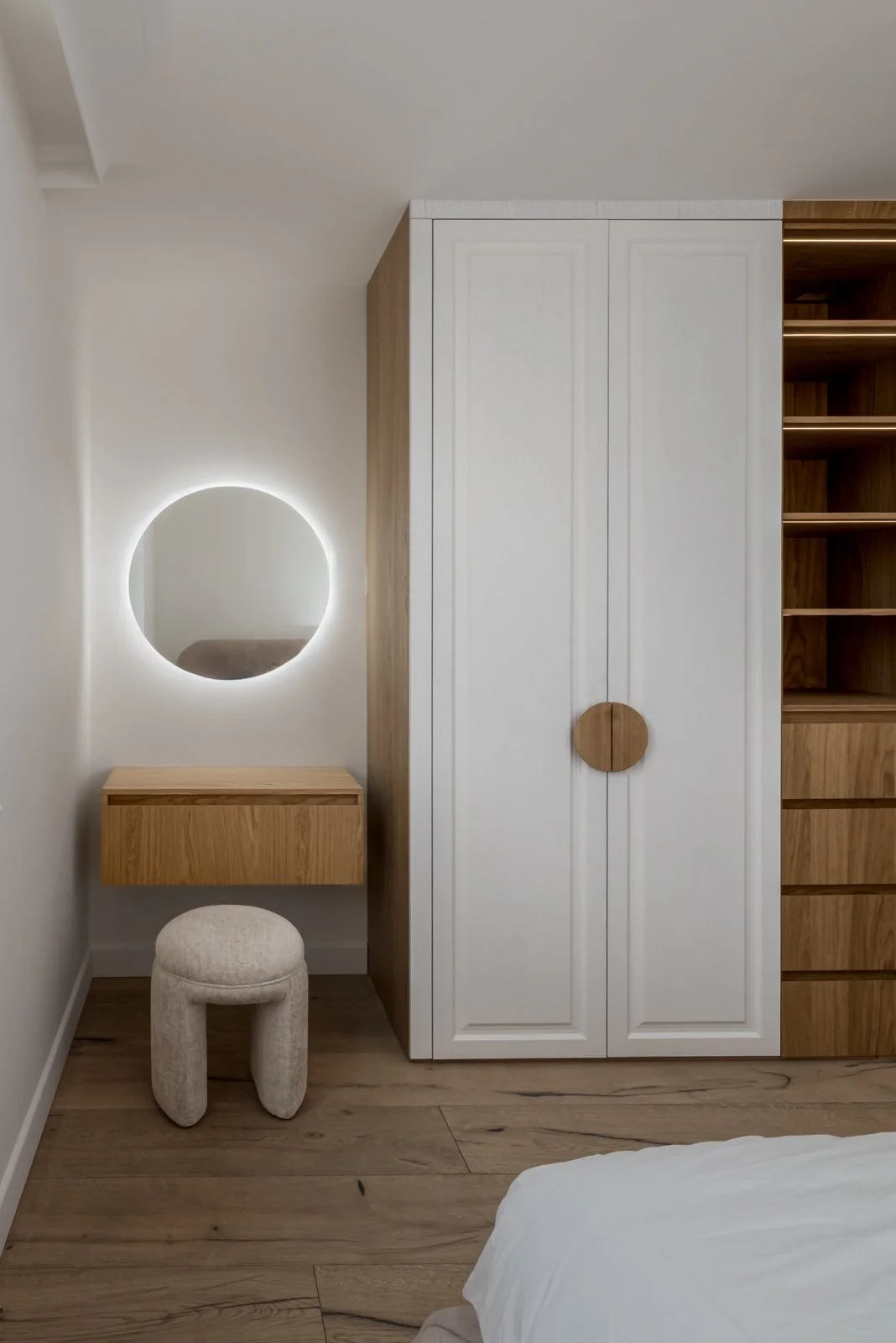 Modern bedroom corner with white and wooden wardrobe, round mirror with LED lighting, small wooden vanity, beige cushioned stool, and part of a white bedsheet visible in the lower right corner.