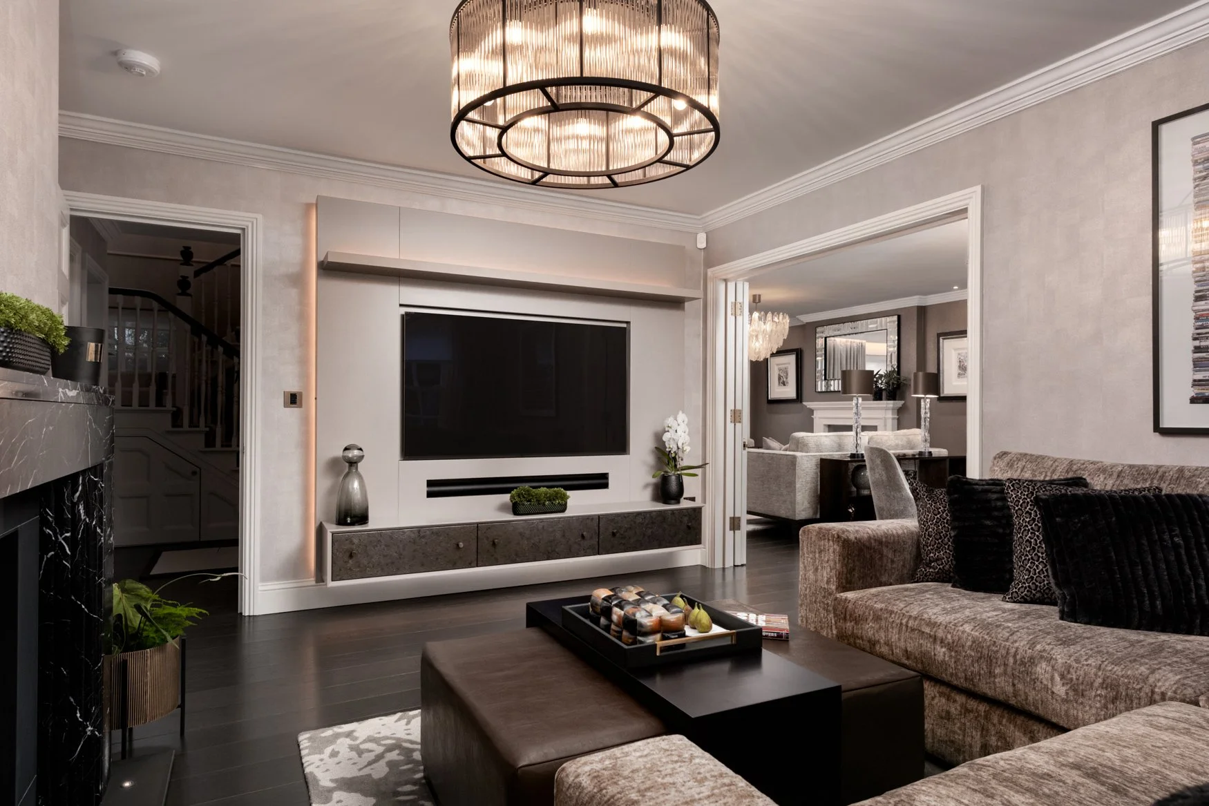Living room with a large black flat-screen TV, a modern chandelier, beige sofa with patterned pillows, coffee table with decorative objects, and a partial view of the adjacent room with a chandelier, fireplace, framed art, and additional seating.