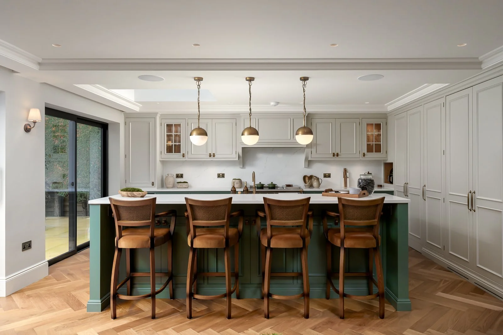 Modern kitchen with white cabinets, a large island with a green base, and five wooden barstools. The kitchen has pendant lights hanging above the island, a sliding glass door on the left, and hardwood floors.