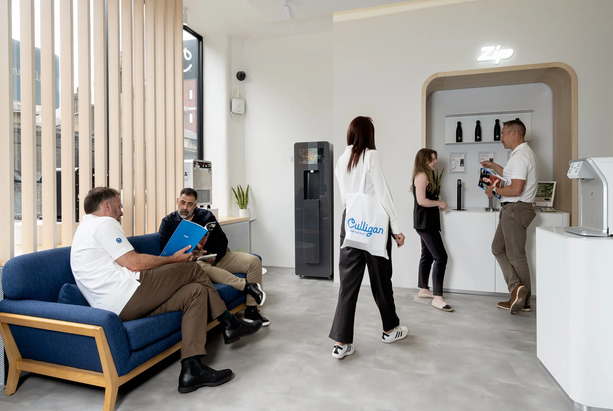 A waiting area and reception at Zip, a business or service location, with four people visible: two men and two women, engaged in different activities inside the modern, clean interior.