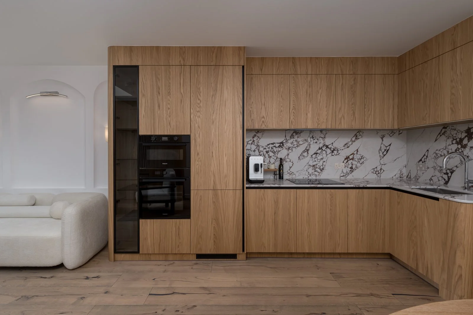 Modern kitchen with wooden cabinets, marble backsplash, and built-in appliances. Part of a living room with a white sofa visible on the left.