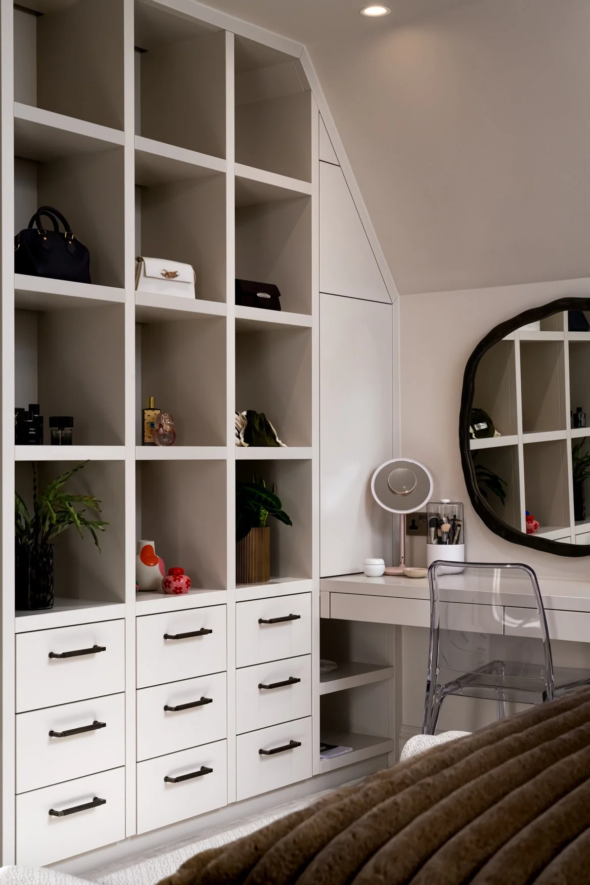 White shelving unit with handbags, perfume, plants, and decorative items in a bedroom or dressing room, next to a vanity with a mirror and makeup brushes.