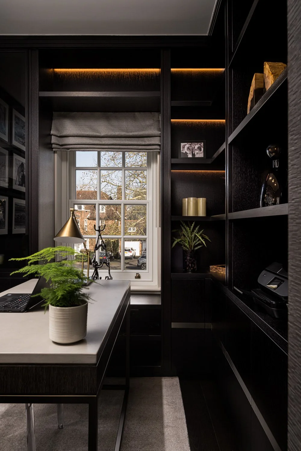 A modern home office with a window revealing a street view, decorated with plants, a laptop, and a gold desk lamp, surrounded by dark wood shelving with decorative items.