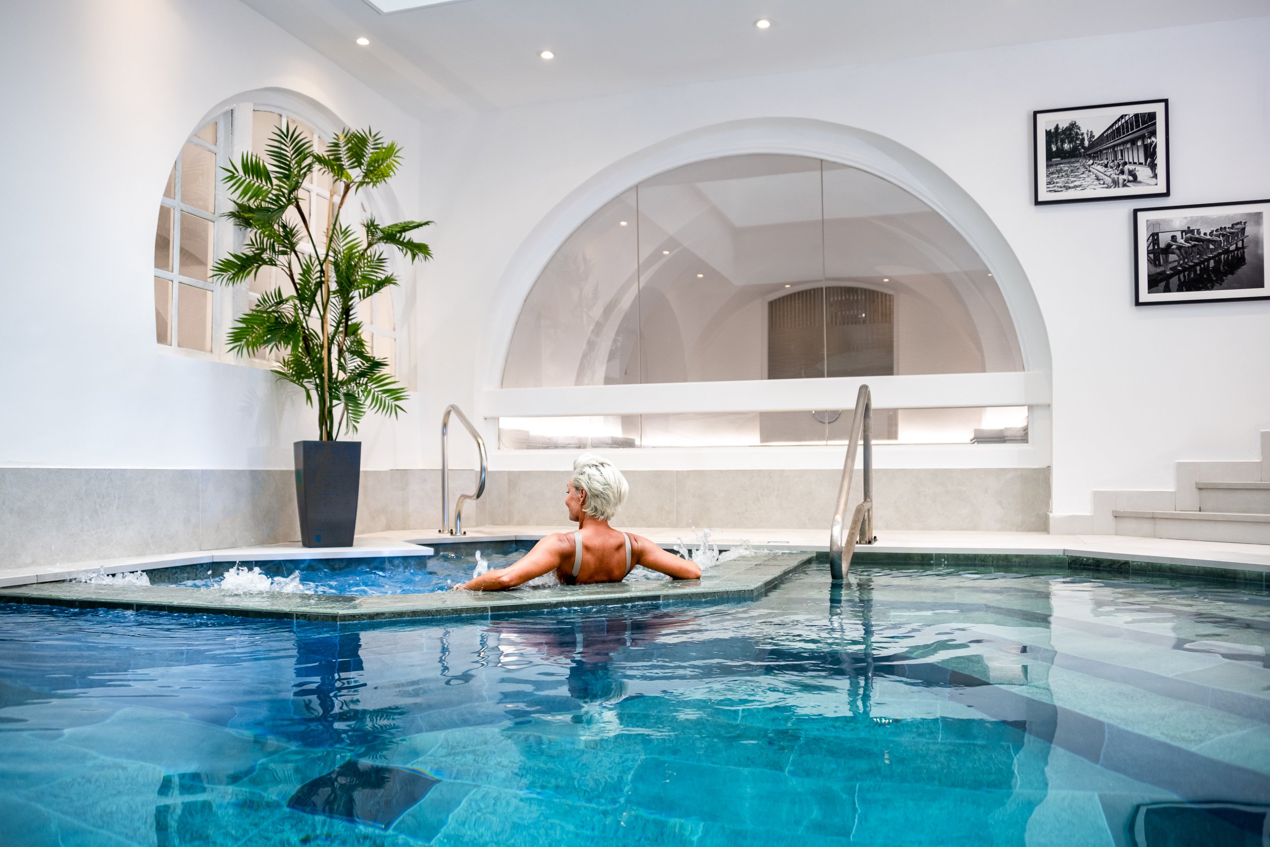 An elderly woman relaxing in a hot tub or spa inside a modern, white-walled building with black-and-white framed photographs and a large potted plant near a window. Gym and Wellness Club in St John  