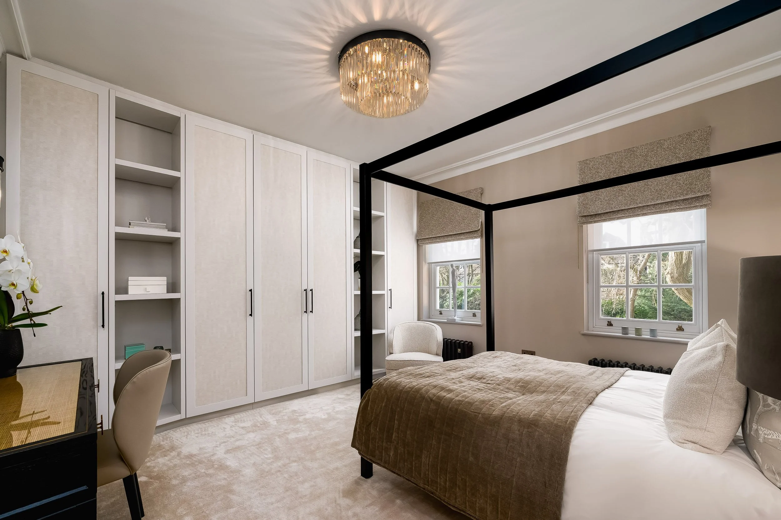 A bedroom with a black four-poster bed, white built-in cabinets, beige walls, two windows with beige shades, and a ceiling chandelier with crystal accents.