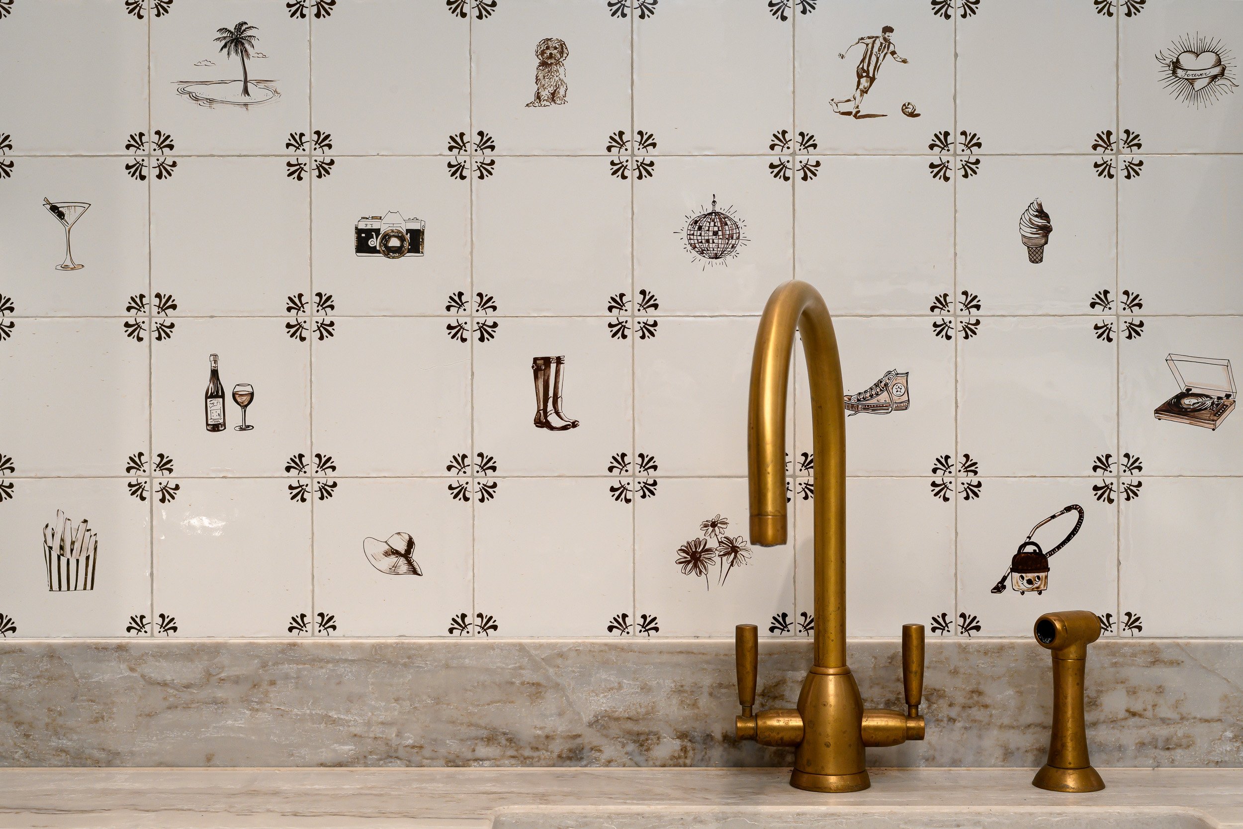 A kitchen detail photographed in London, featuring a brass tap, marble worktop and hand-painted illustrated wall tiles. The playful tile drawings add personality, while the aged brass and marble bring warmth, texture and a refined material finish to 