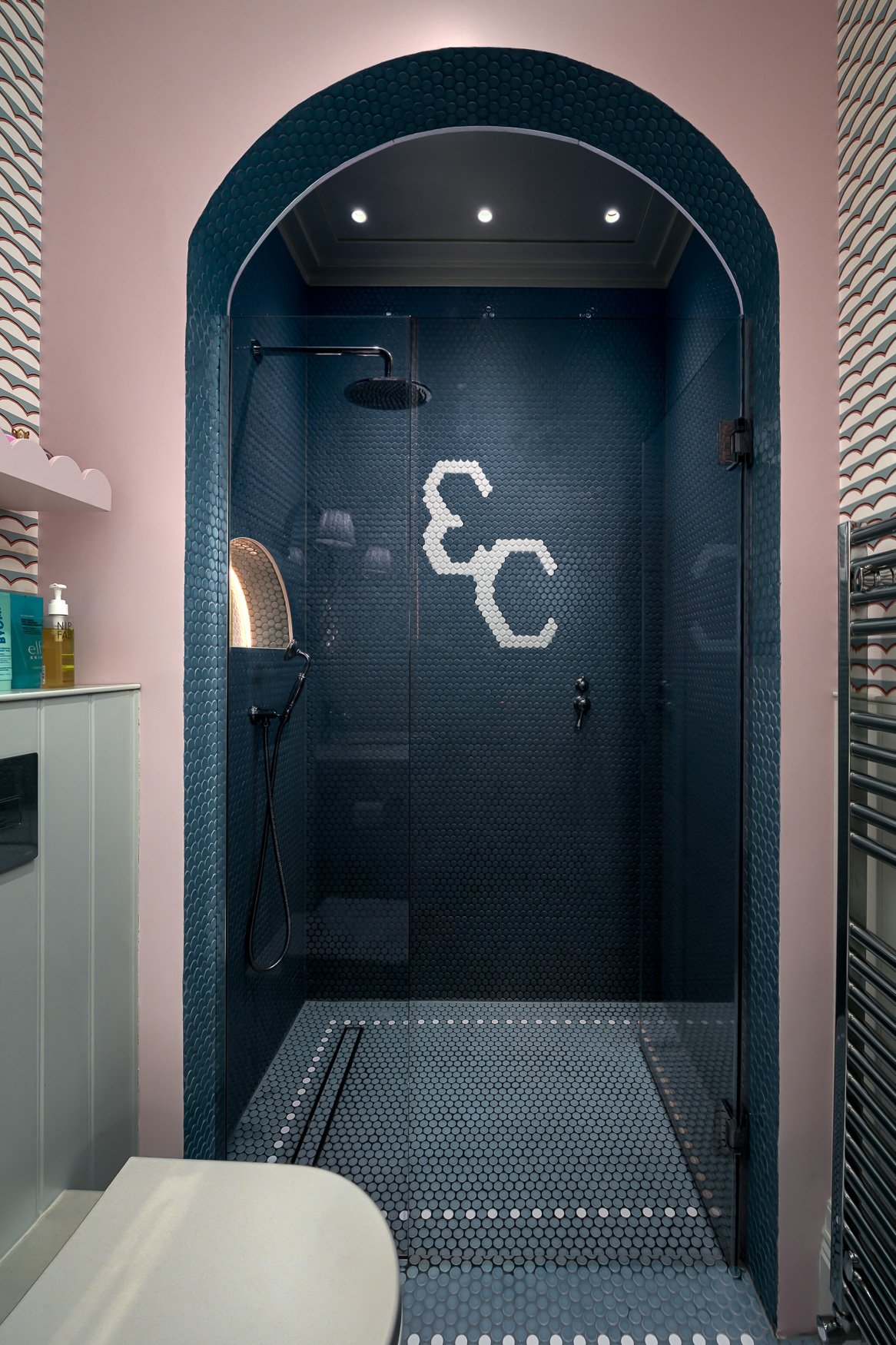 Children’s bathroom with blue tiled shower arch, pink walls and playful patterned wallpaper.