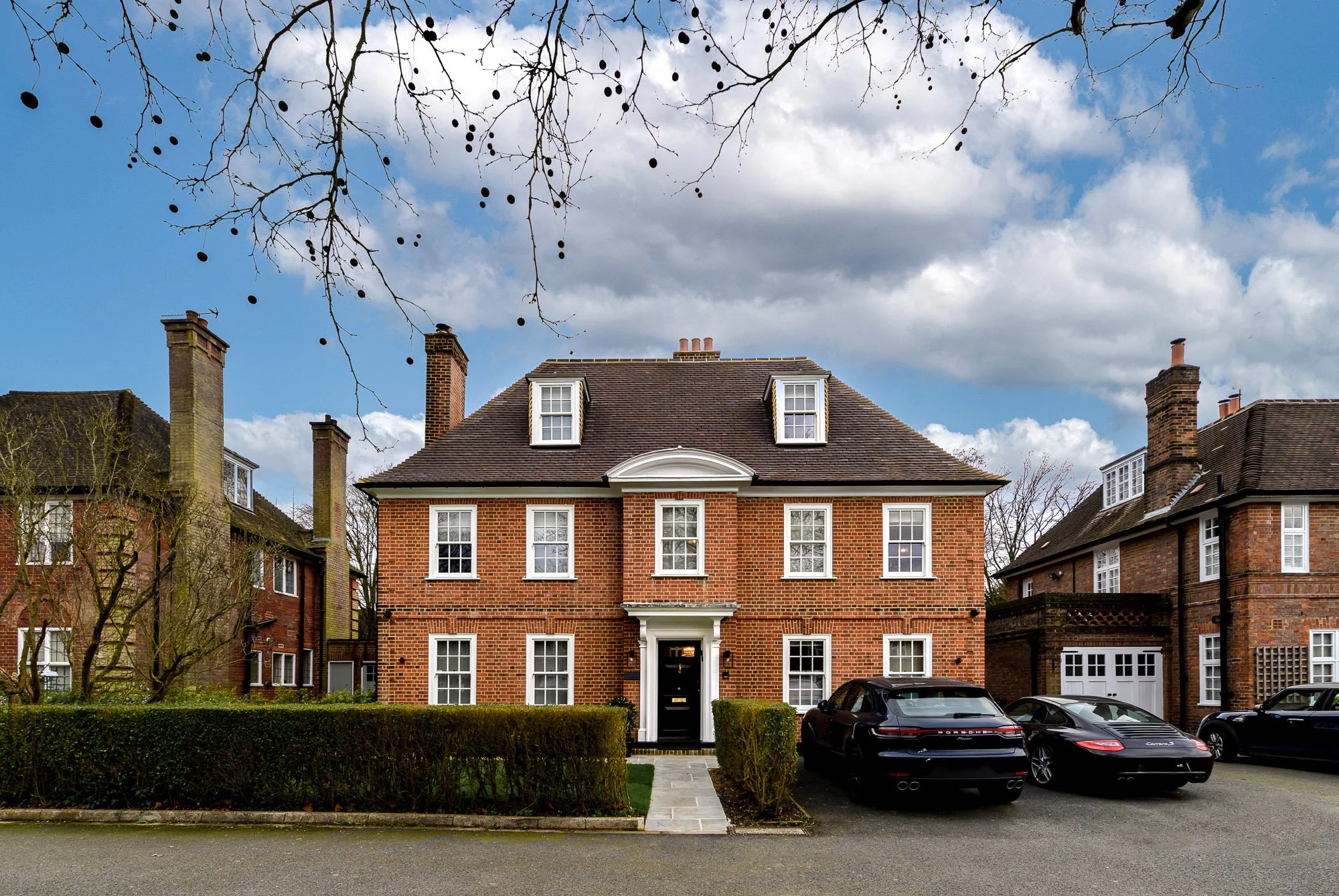Exterior architectural photograph of a large brick residential property with symmetrical facade, dormer windows, front entrance, driveway and surrounding period homes.