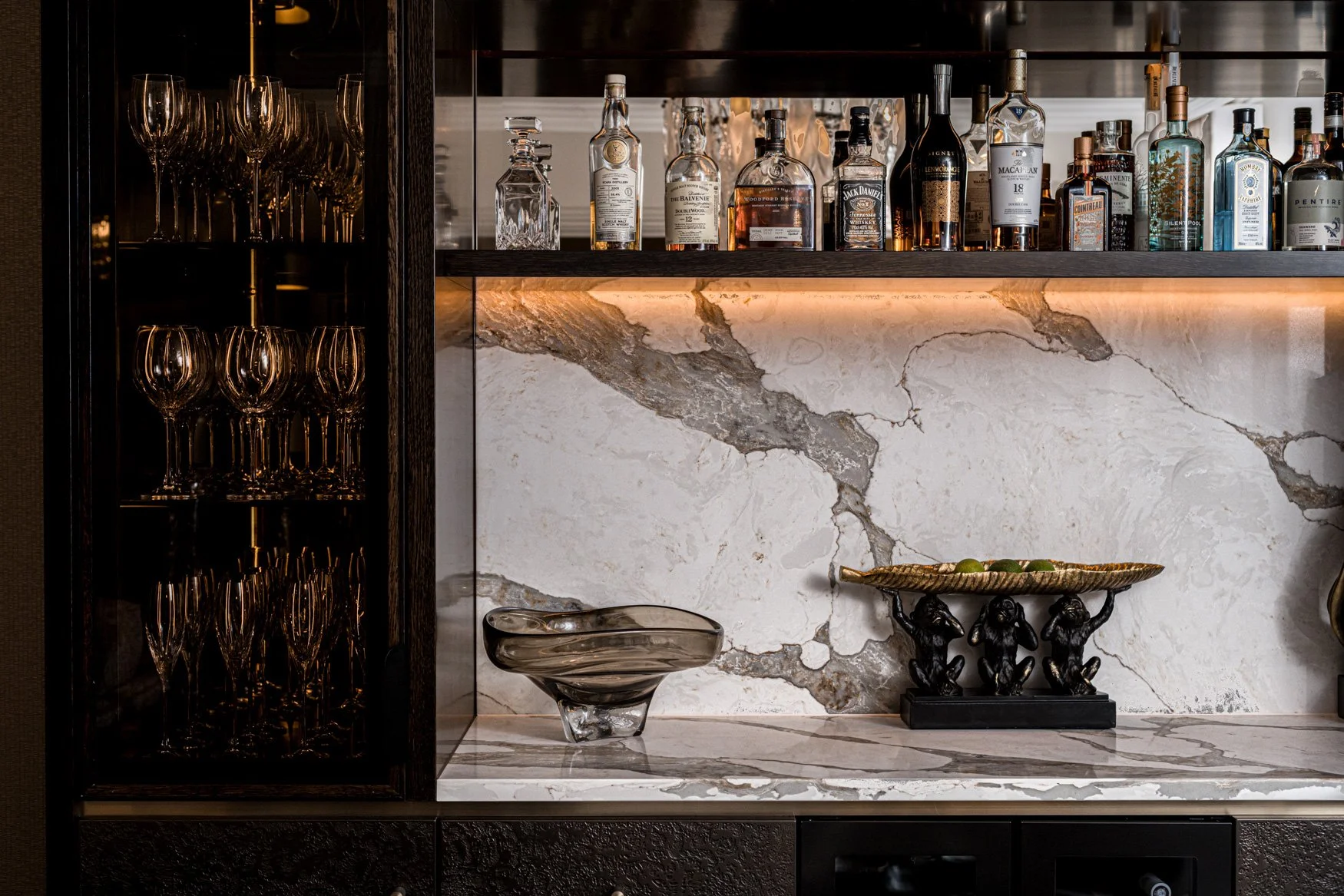 A luxury home bar photographed in London, featuring dark bespoke joinery, glassware, backlit shelving, a marble splashback, premium drinks display and carefully styled decorative pieces. The image captures the richness of the materials, the mood of t