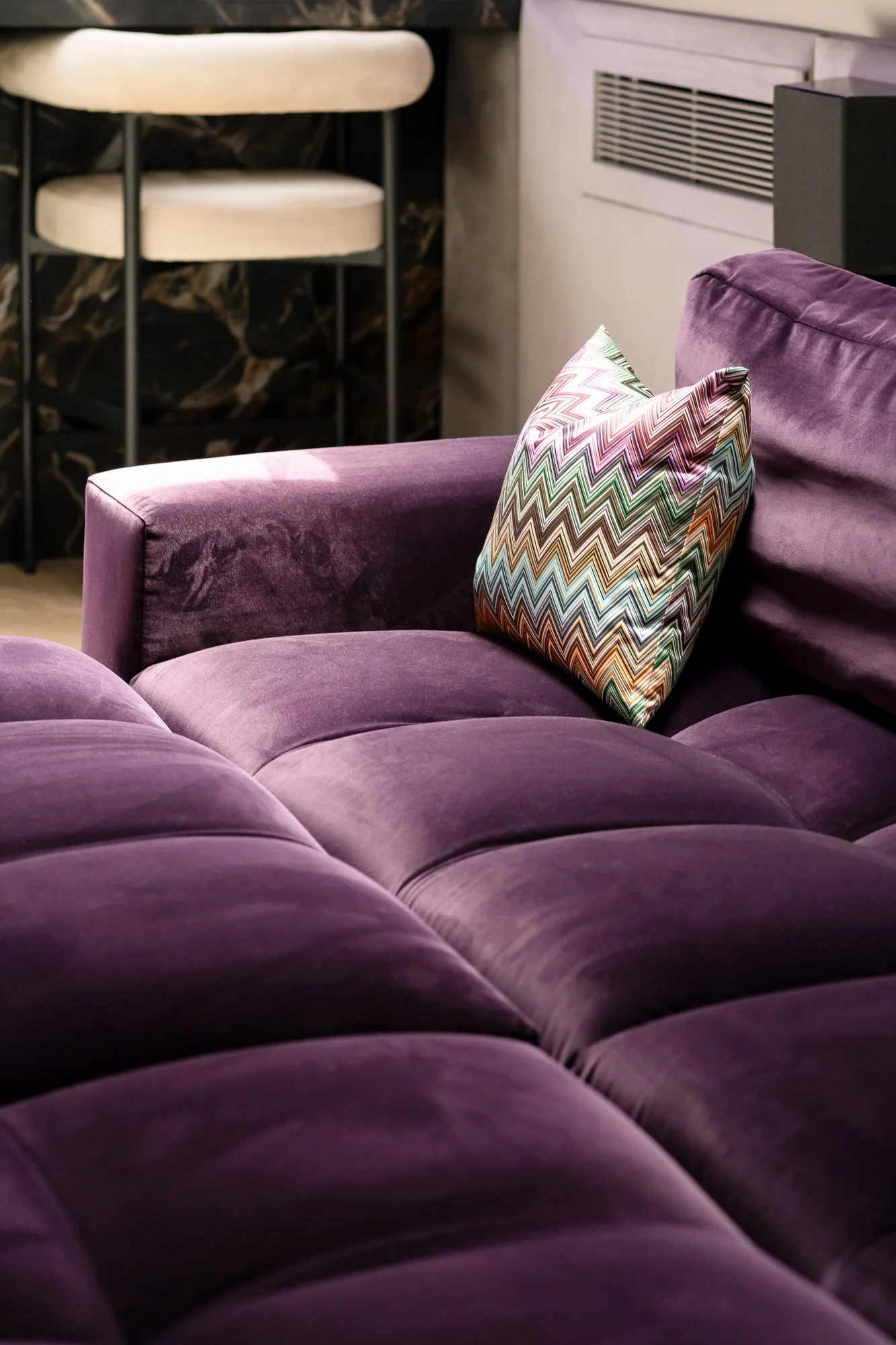 A purple velvet sofa with a multicolored zigzag patterned pillow on it. In the background, there are two white chairs with black legs, and a wall heater or vent on the wall.