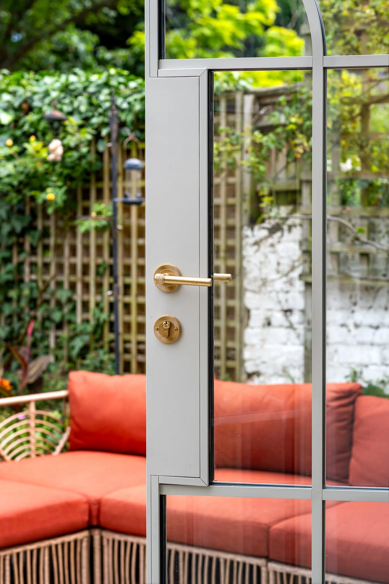 A close-up detail of an arched glazed garden door with a brass handle and slim metal frame. The image shows the door finish, ironmongery and glazing detail, with soft garden seating and greenery visible through the glass.