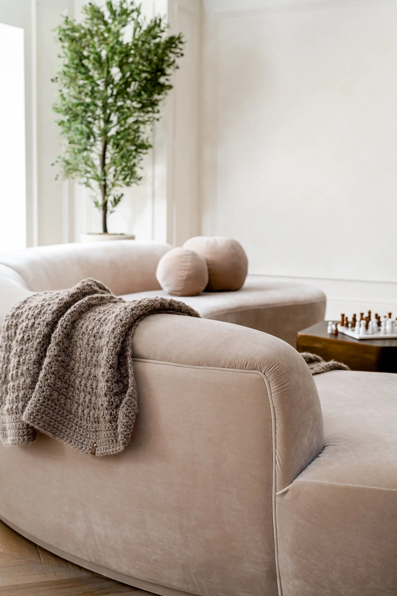 A beige curved sofa with a fuzzy gray throw blanket draped over it, next to round beige bolster pillows, in a bright living room with white walls, a tall potted plant, and a wooden chessboard on a coffee table.