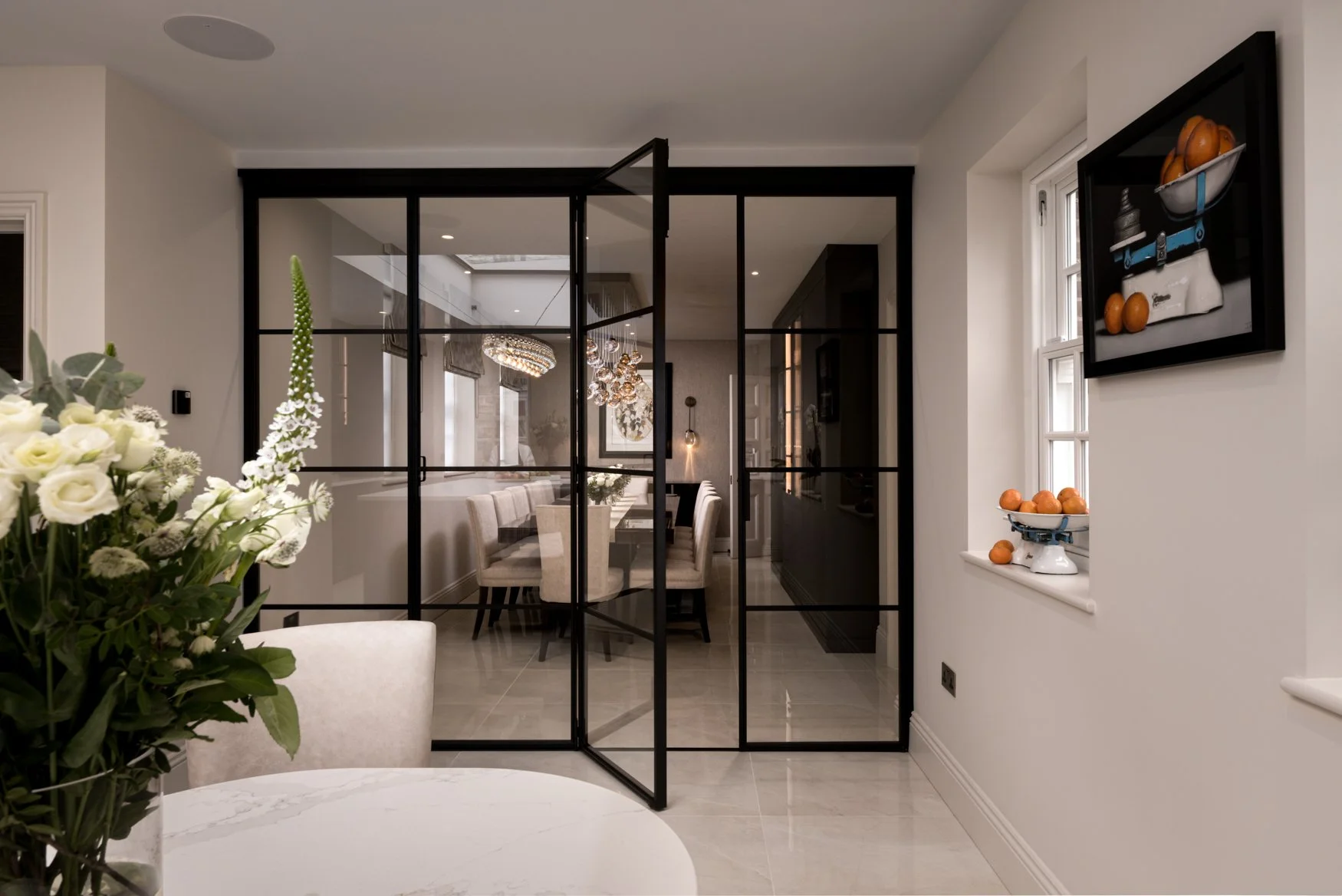 An open-plan London interior photographed through black steel-framed glass doors, showing the connection between kitchen, dining and living spaces. The image features soft neutral finishes, marble surfaces, floral styling, framed artwork and architec