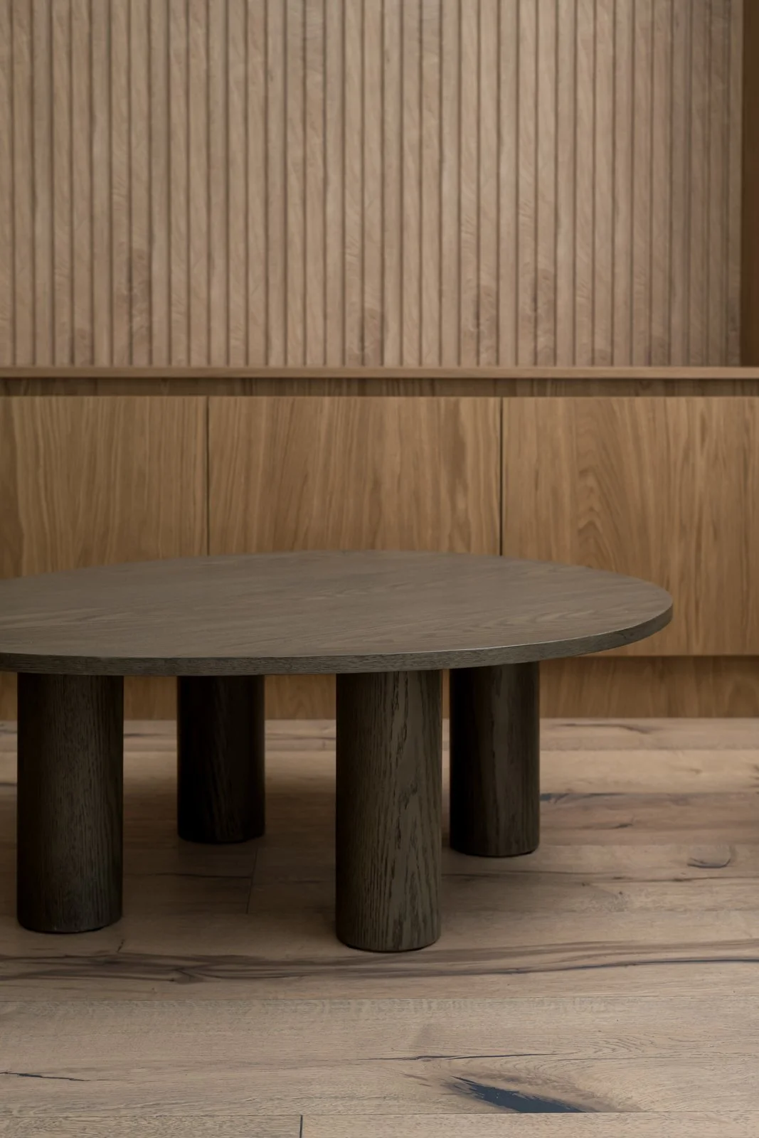 A wooden, round coffee table with dark wooden legs in front of a wooden-paneled wall and wooden floor.