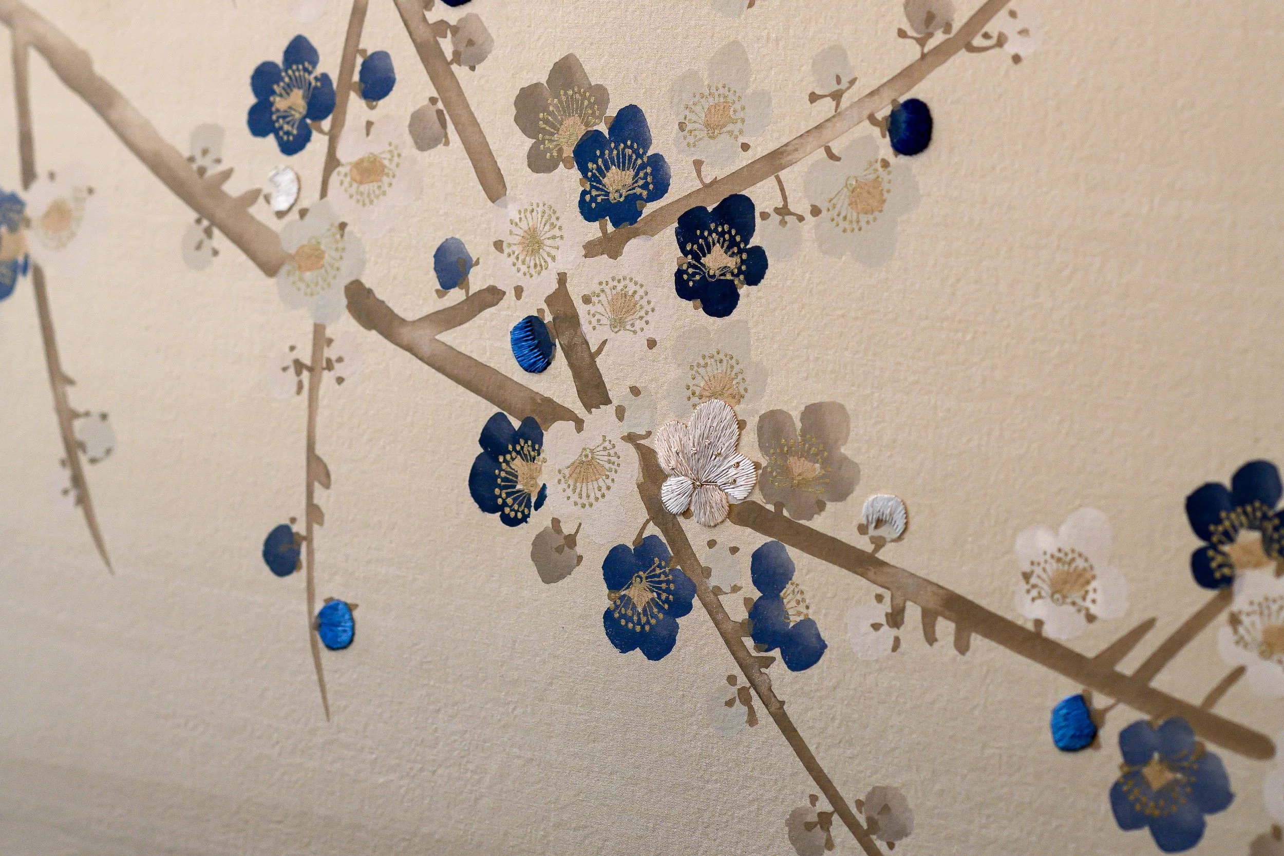 A close-up detail of hand-painted floral wallpaper photographed in London, featuring blue, white and soft neutral blossoms with delicate branch patterns and textured surface detail. The image highlights the craftsmanship, colour palette and quiet dec