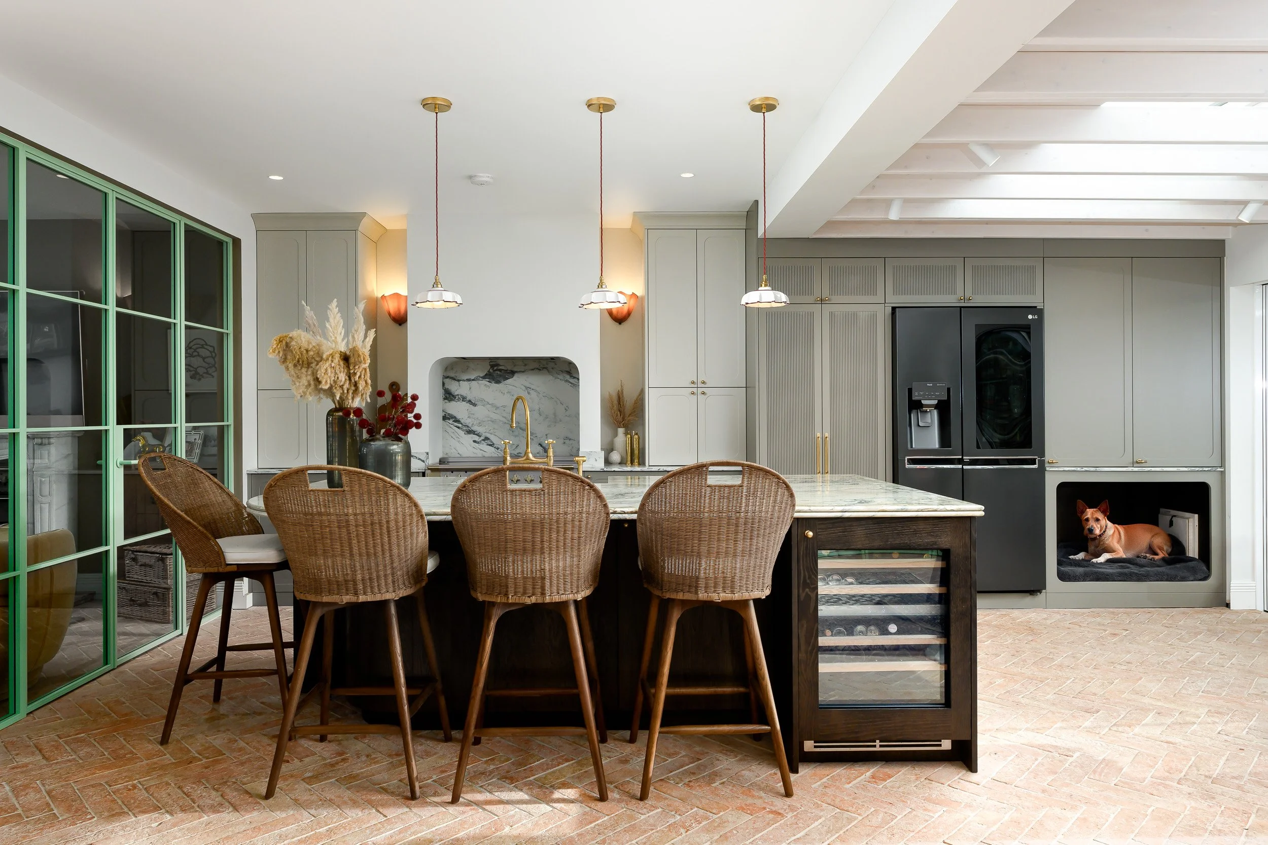 A spacious bespoke kitchen photographed in London, featuring green-framed glass doors, a large marble-topped island, woven bar stools, soft neutral cabinetry, brass details, pendant lighting, brick flooring and a built-in dog bed. The image shows a w