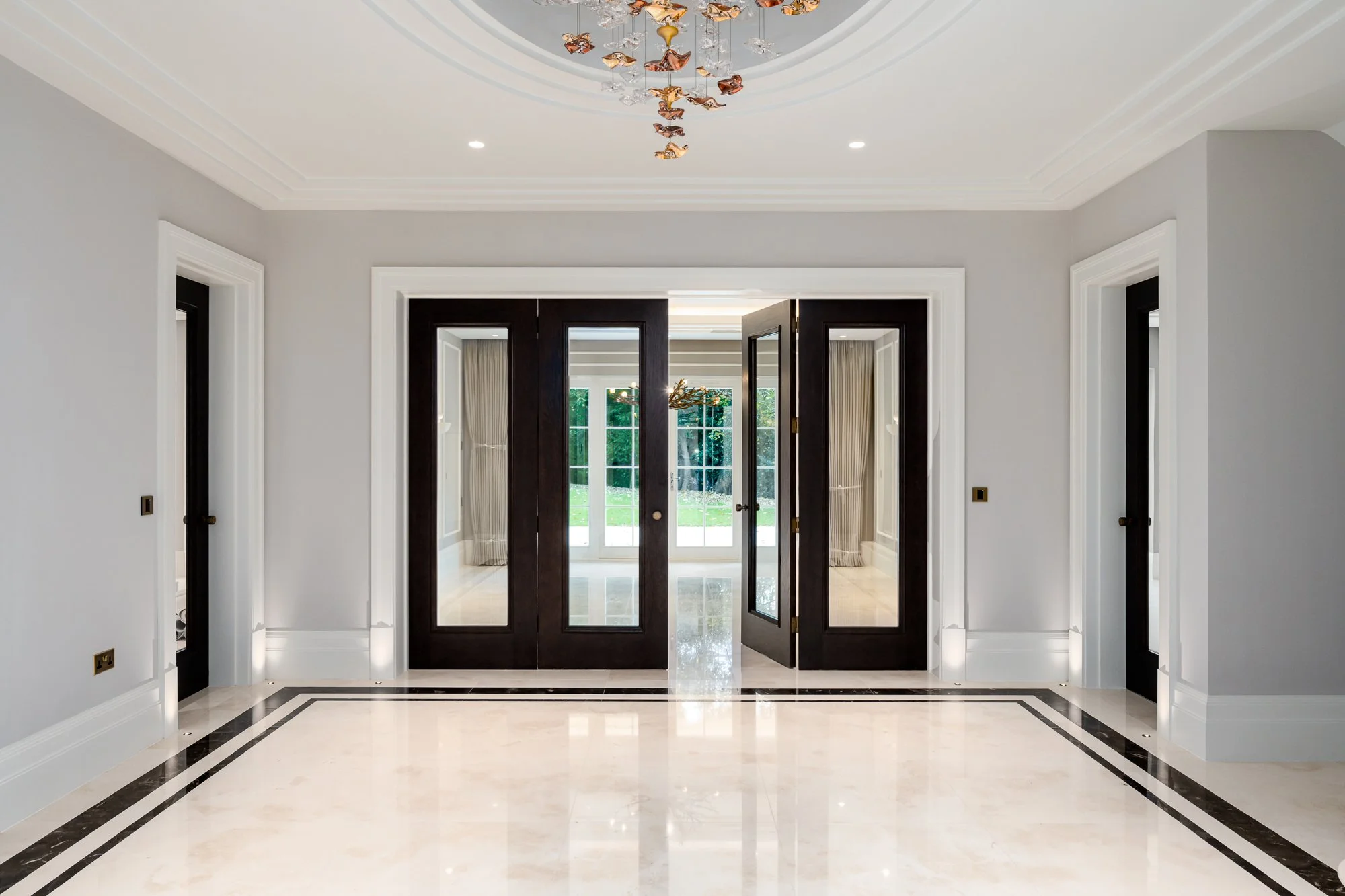 A luxury hallway with dark glazed double doors opening into a bright interior beyond. The image highlights the scale of the doors, the dark timber finish, glass panels, polished floor and formal architectural detailing around the entrance.