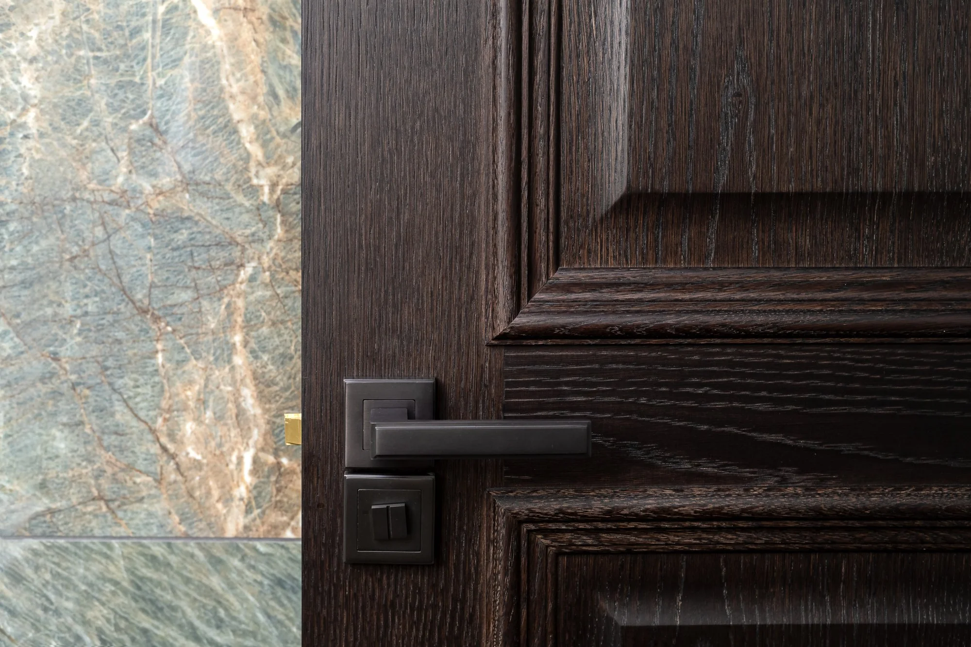 A close-up detail of a dark timber interior door with a black handle, set beside marble wall finishes. The image highlights the wood grain, panel moulding, hardware choice and material contrast.