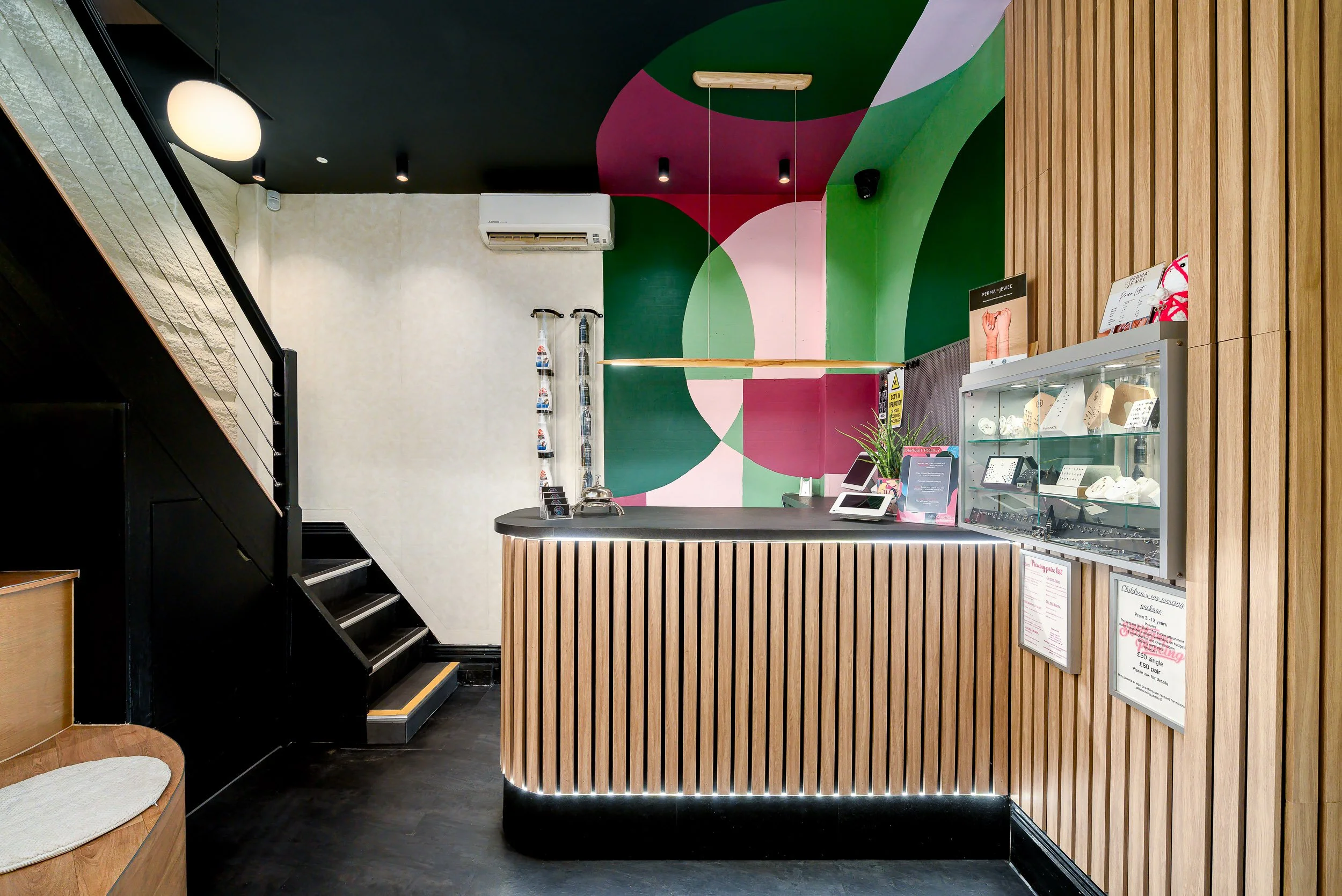 Interior of a tattoo studio in London  with a wooden curved counter, colorful abstract wall mural, display case with jewelry, and decorative lighting.