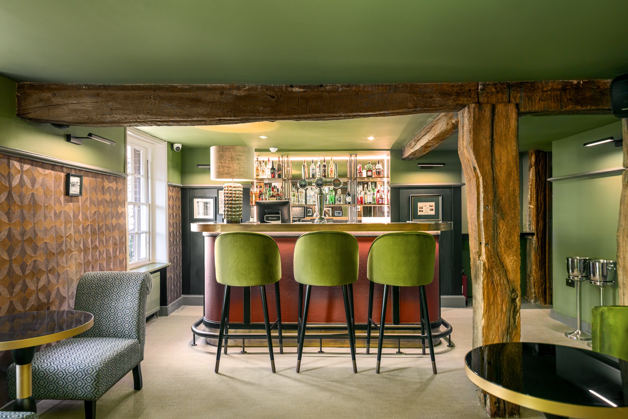 Interior of a bar with green chairs, a bar counter, and a well-stocked liquor shelf behind it. Wooden beams and patterned wallpaper are visible.
