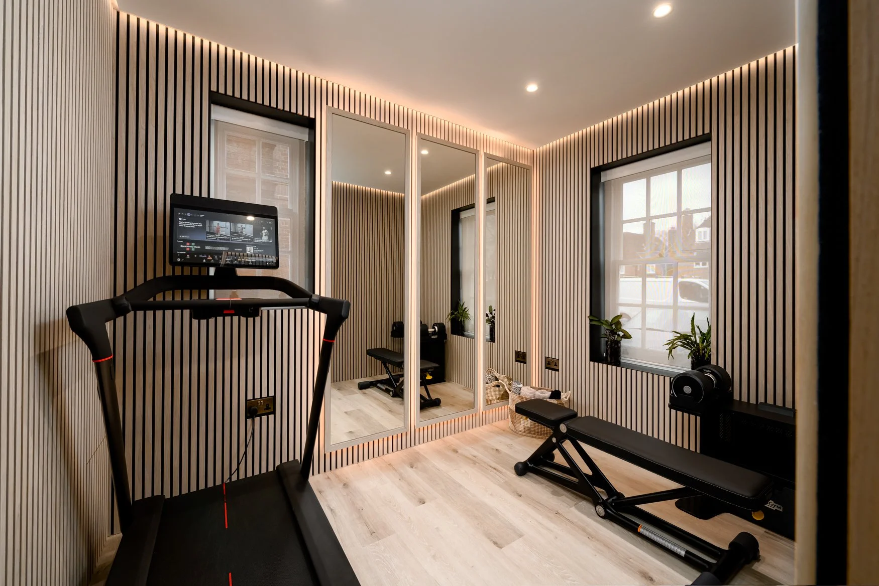 Home gym with treadmill, weight bench, and mirror, featuring wood-paneled walls, a window with a view, and indoor plants.