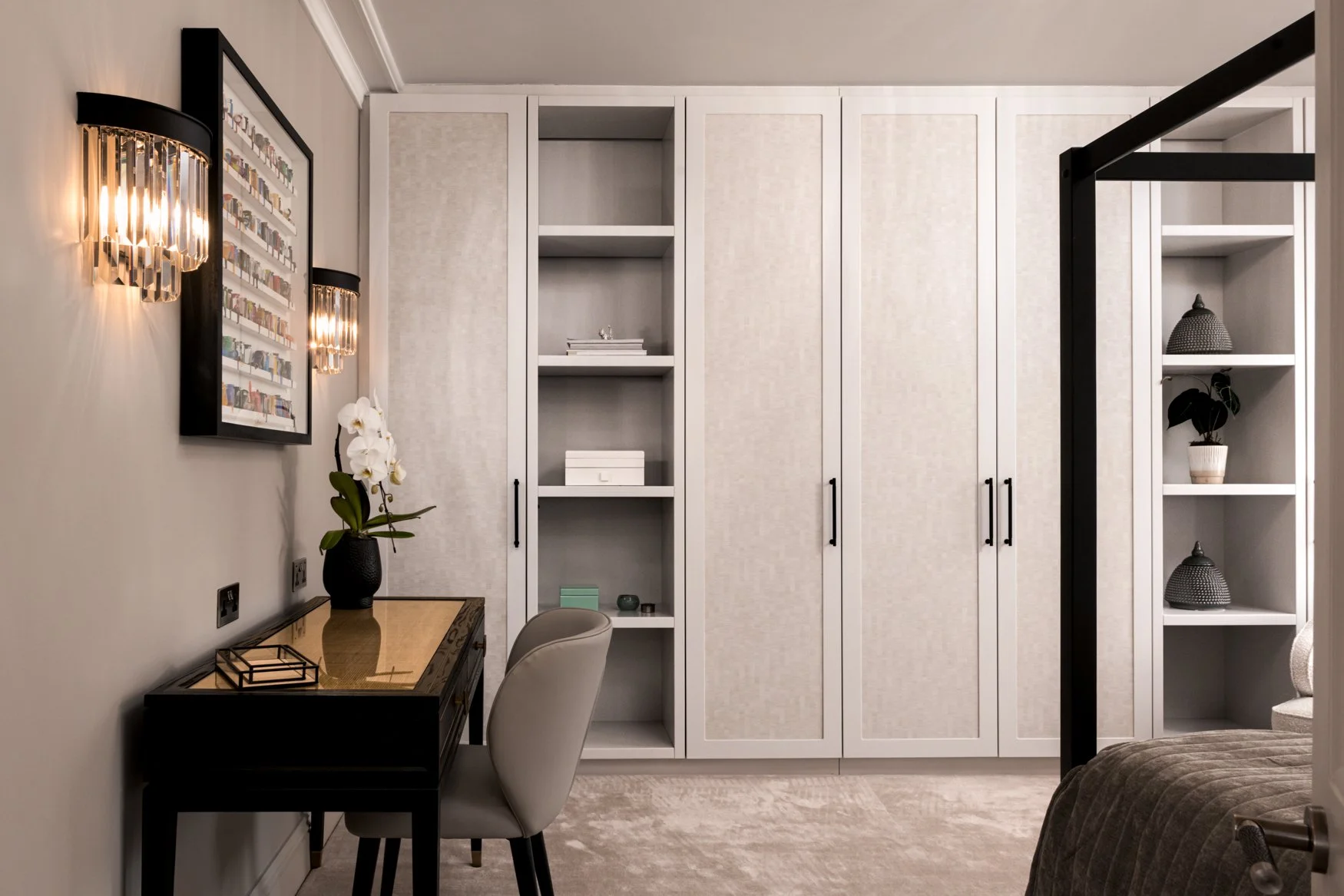 A modern bedroom corner with white built-in cabinets, a black and gold desk, a white chair, a wall-mounted light with crystal accents, a framed artwork of books, a black vase with a white orchid, various decorative items on the shelves, and part of a