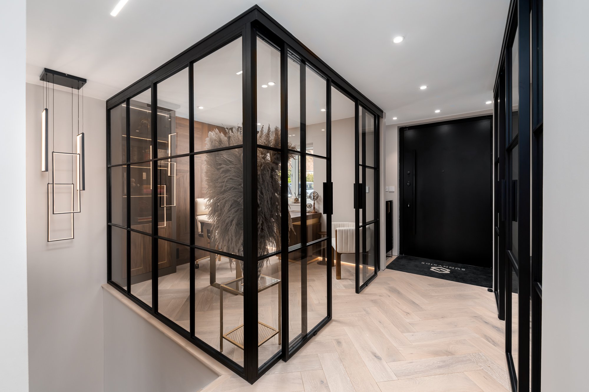 A modern hallway with a black framed glass partition enclosing a luxury home office. The image highlights the slim metal glazing, full-height glass panels and open doorway, showing how internal glazing can create a separate workspace while keeping th