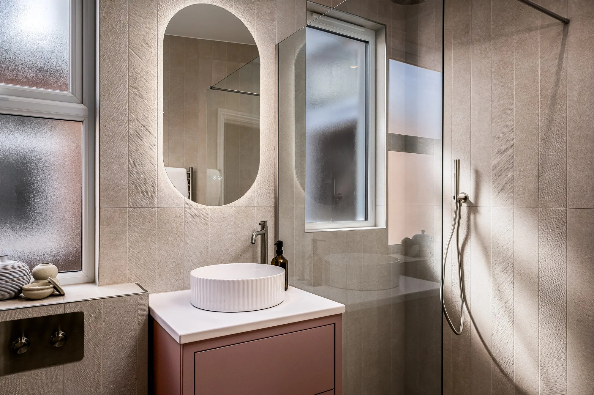 Designed bathroom with pink vanity, arched mirror, textured finishes and natural light from the window.
