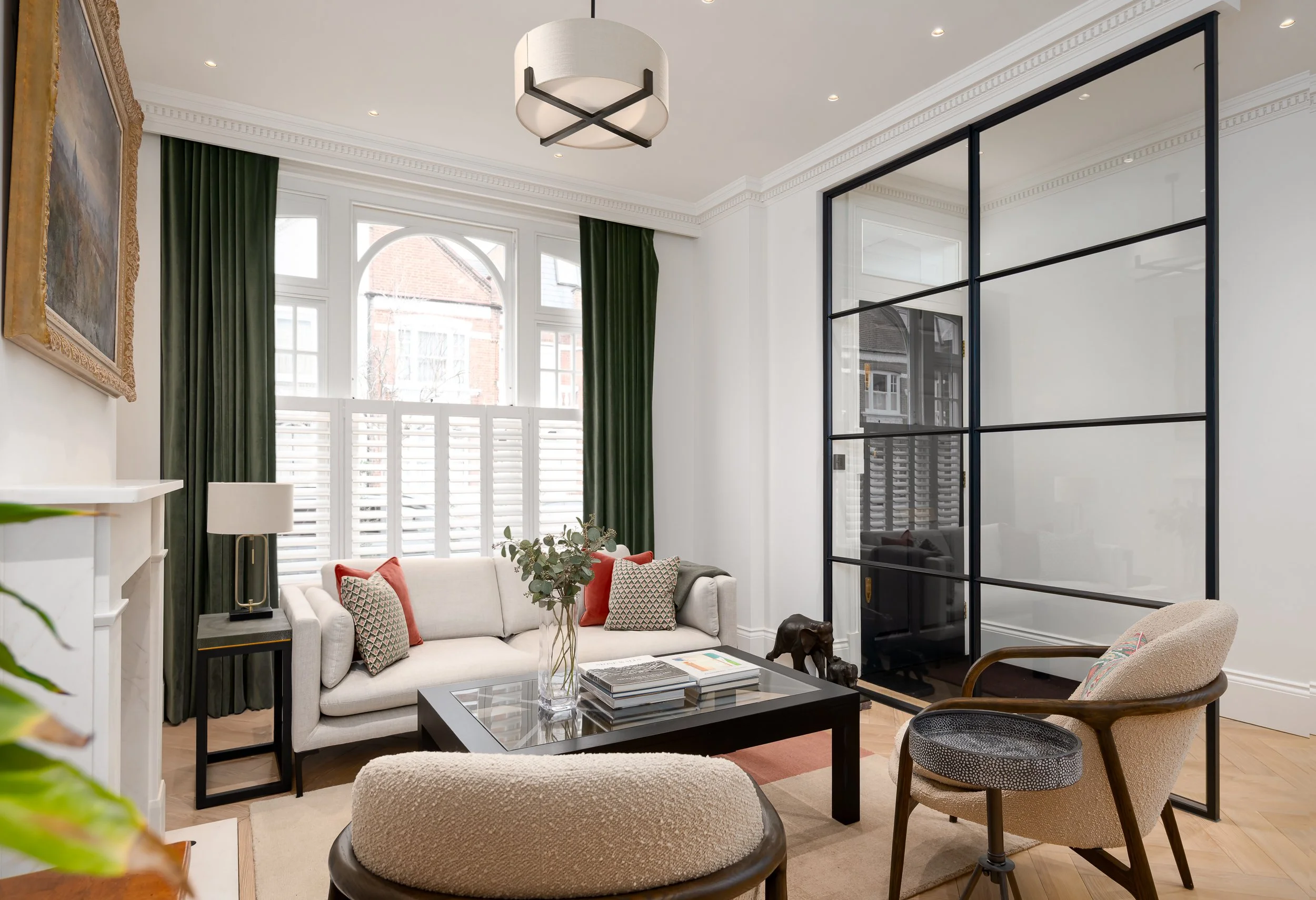 A bright London living room photographed with black-framed internal glazing, green curtains, soft neutral seating, layered cushions, timber flooring and considered styling. The image shows the balance between architectural structure and comfortable i