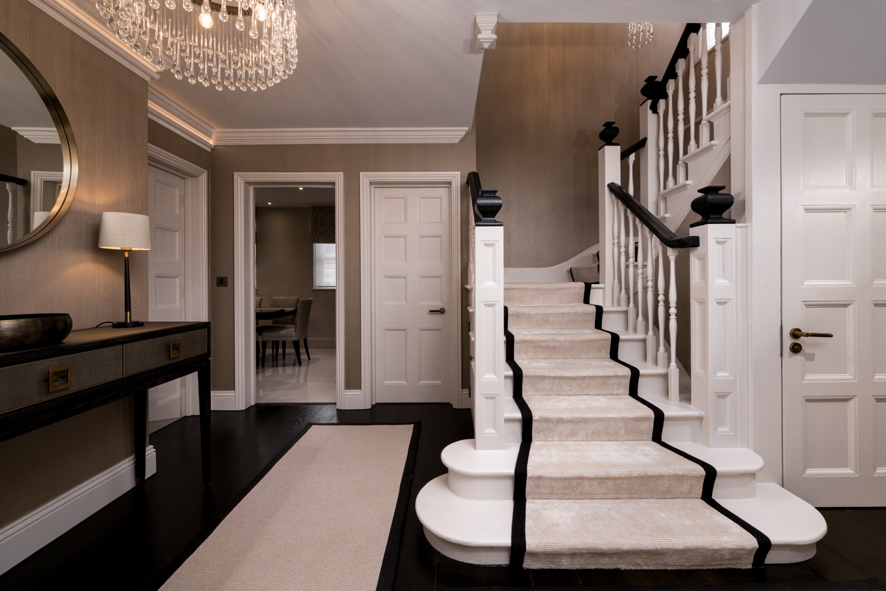 A luxury entrance hall photographed in London, featuring a sweeping staircase with a black-bordered runner, crystal chandelier, warm wall finishes, console table, mirror, layered lighting and classic architectural detailing. The image captures the se