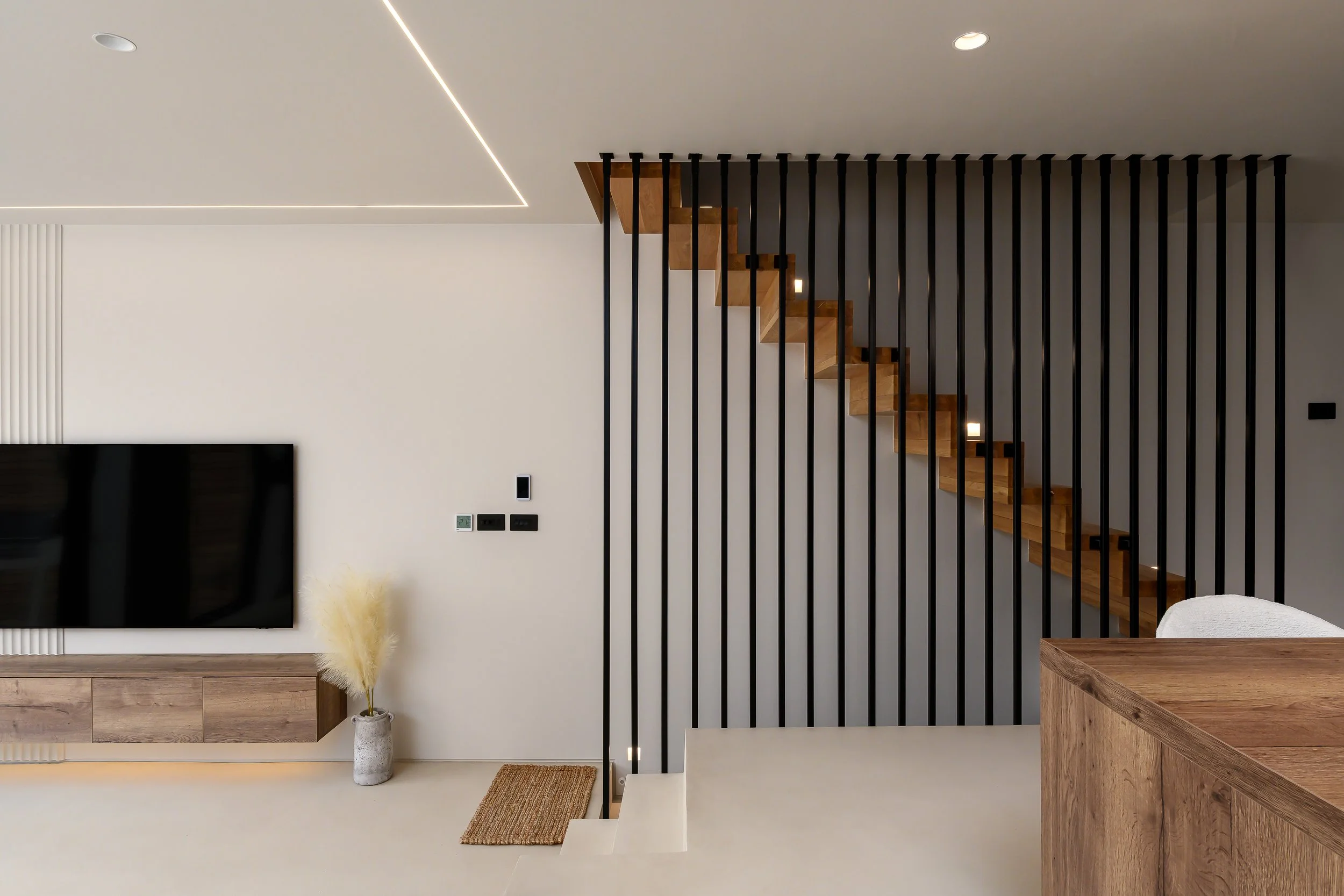 Interior architecture photograph of a modern residential staircase with timber treads, black vertical slatted screen, integrated ceiling lighting and contemporary open-plan living space.