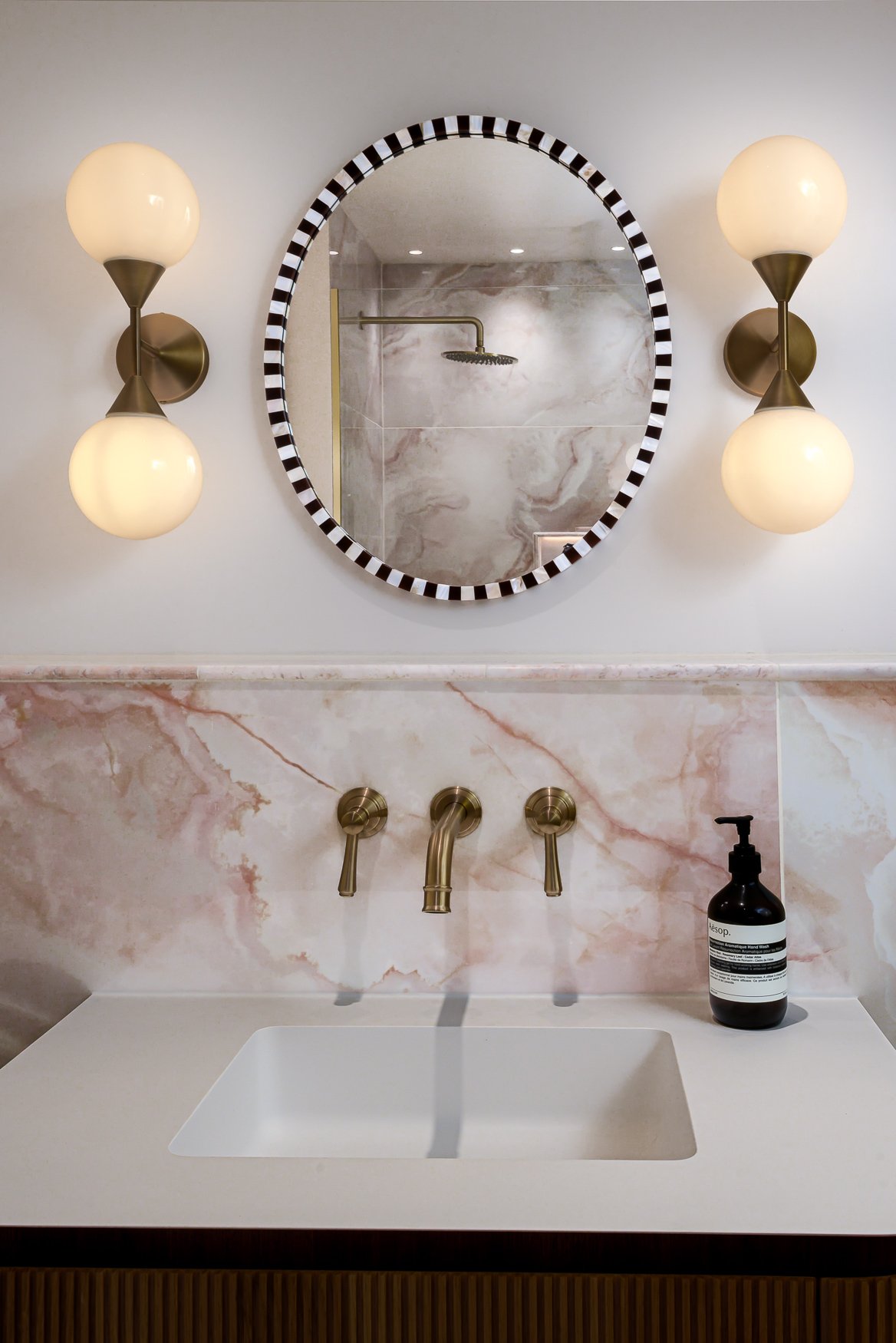 Bathroom vanity with pink marble-effect tiles, brass wall taps, mirror and globe wall lights.