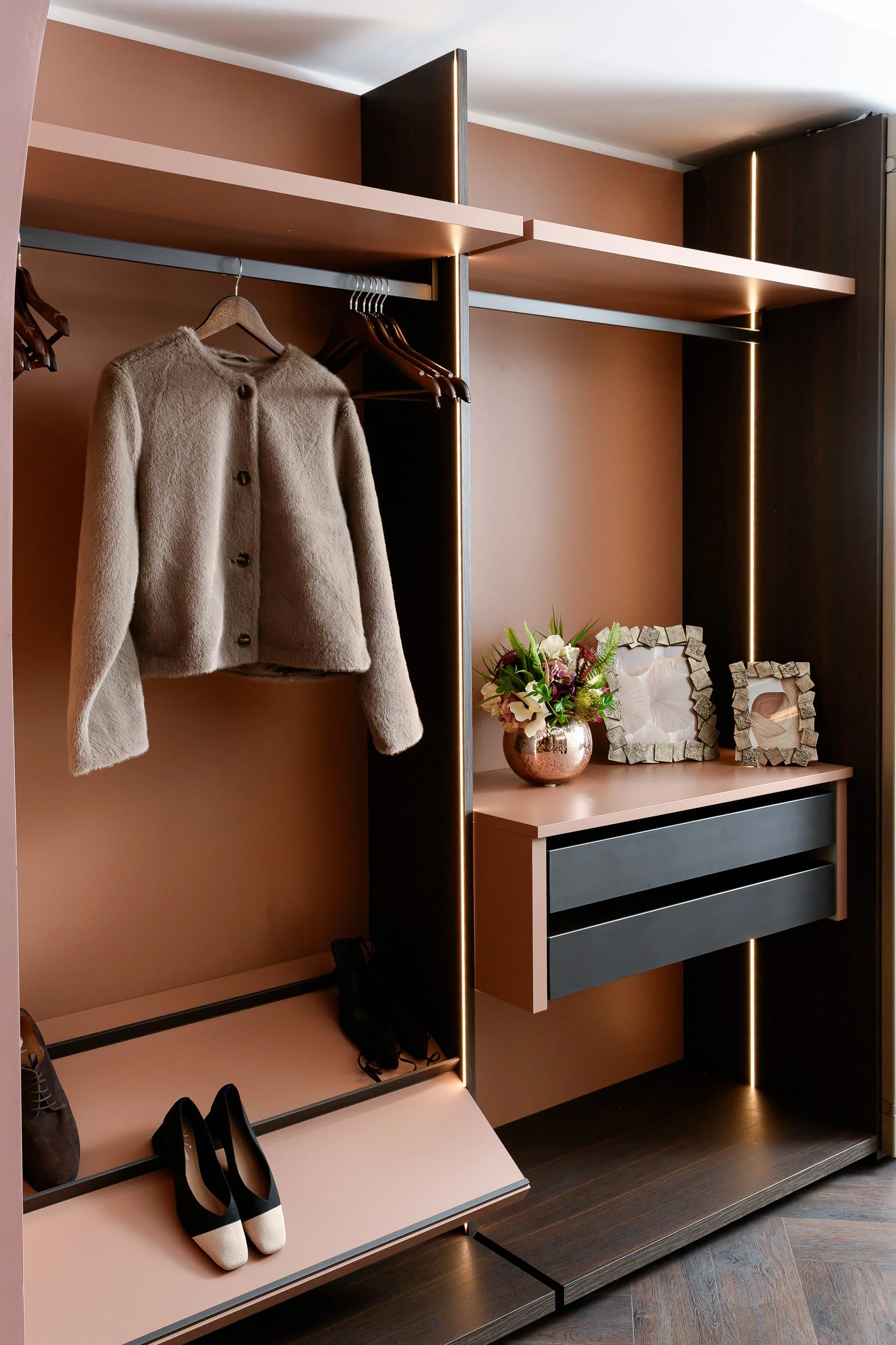 A modern wardrobe with hanging clothes, shoes, and decorative picture frames on a pink and black storage unit. There's a beige coat on a hanger, multiple hangers, a pair of black and white shoes, and a potted flower. Showroom, London.