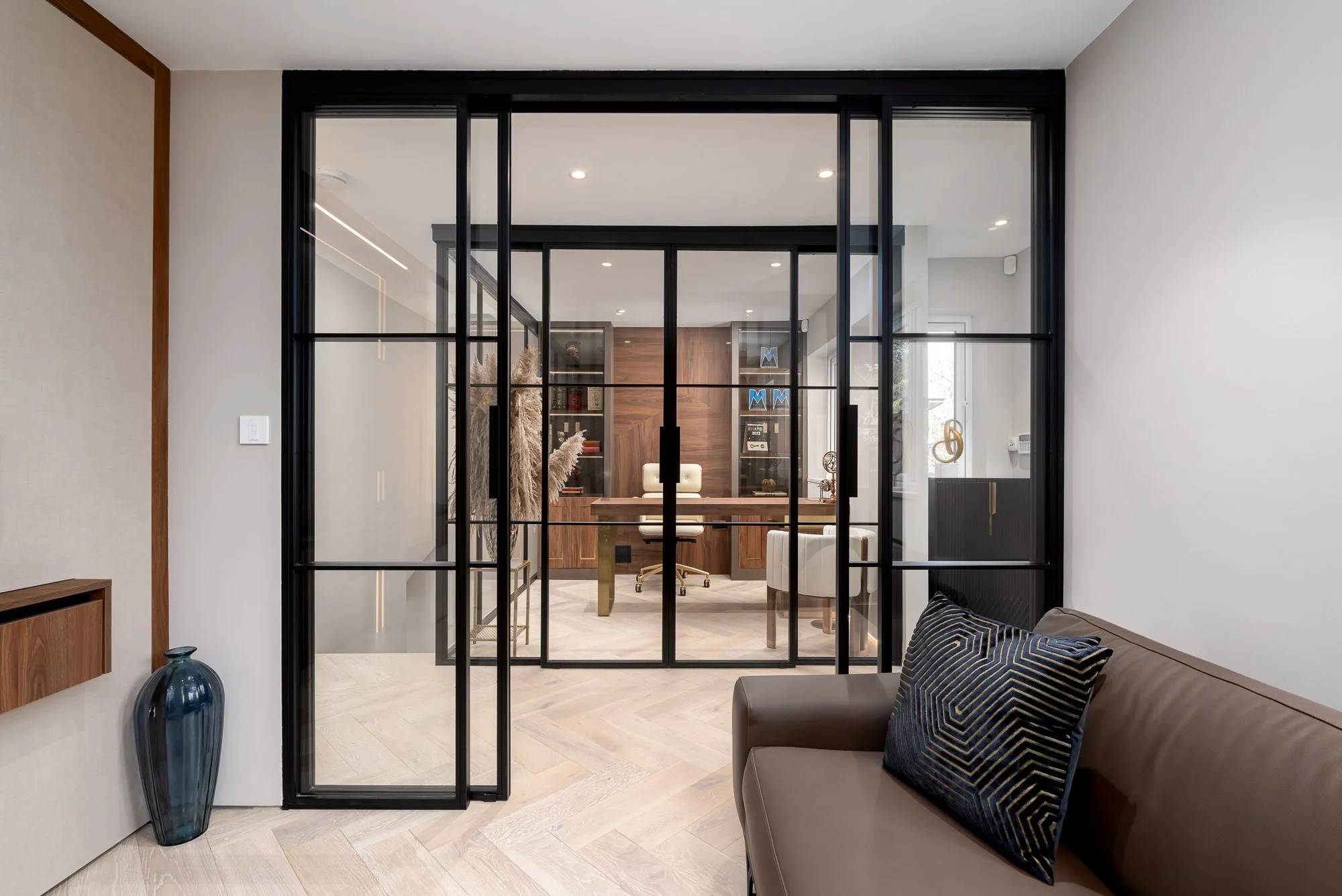 A modern residential interior showing black framed glass doors opening into a home office. The photograph highlights the slim metal frames, glazed panels and how the doors separate the workspace while keeping the room visually connected.