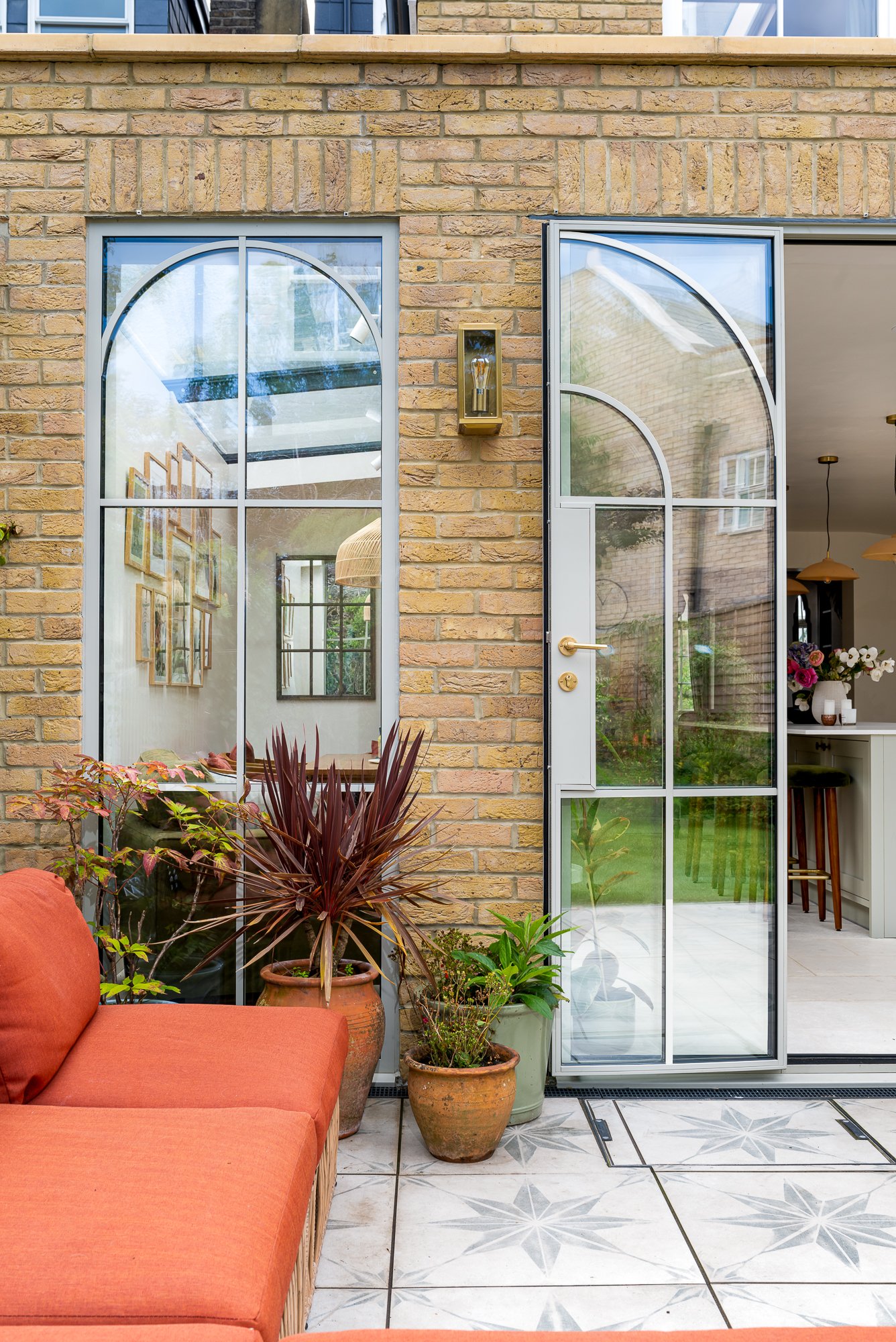 An exterior view of an arched glazed patio door and matching window set into a brick residential extension. The image highlights the curved metal glazing detail, slim frames, brass handle and the connection between the indoor dining space and garden 