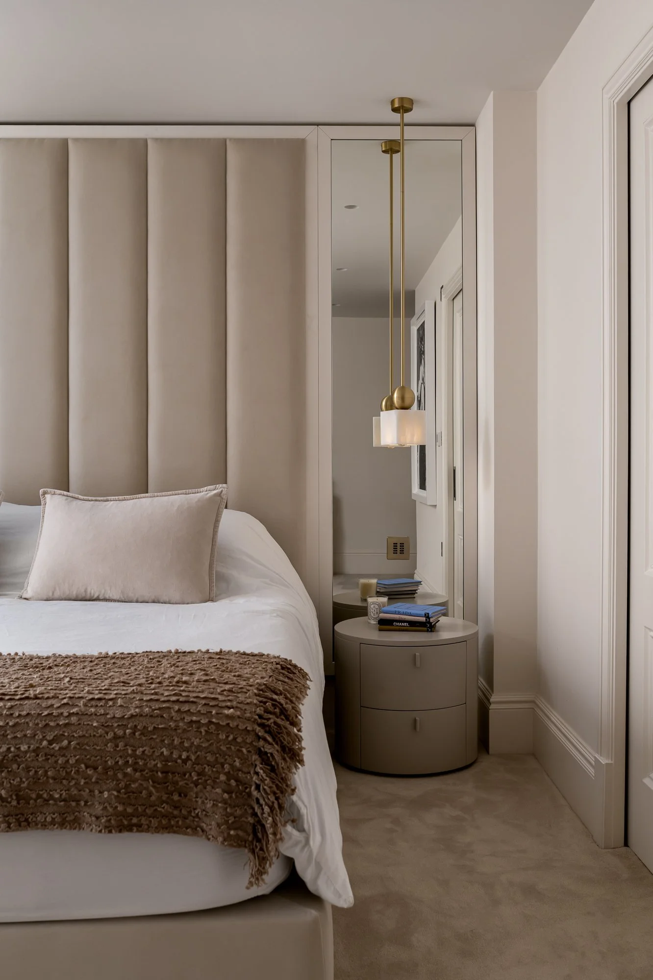 A cozy bedroom corner featuring a white bed with beige pillows and a textured brown blanket, a beige nightstand with books, candles, and a small decorative item, a beige padded headboard, a vertical mirror, white walls, and a modern hanging light fix