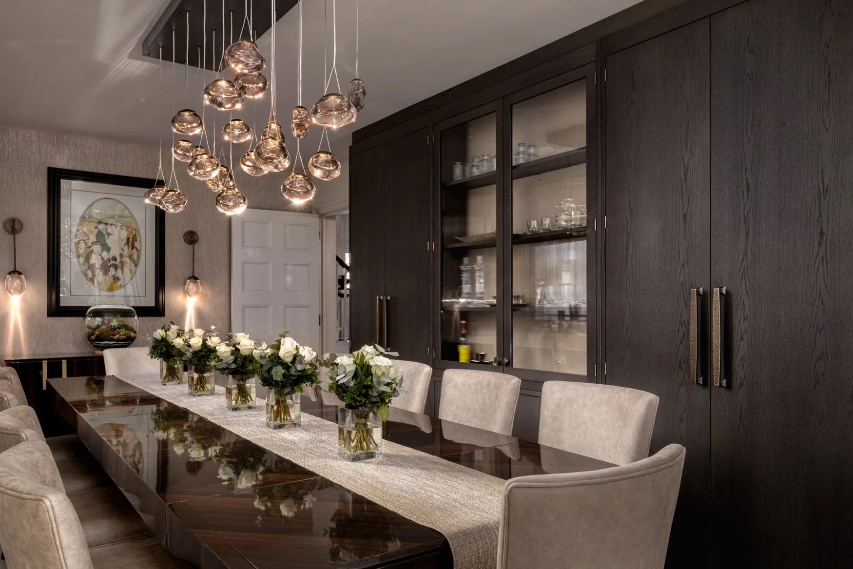 A dining room with a polished wooden table, surrounded by beige upholstered chairs. The table has vases with white roses and greenery. A black cabinet with glass doors holds glassware and dishes. Modern lighting fixtures hang from the ceiling, and a 
