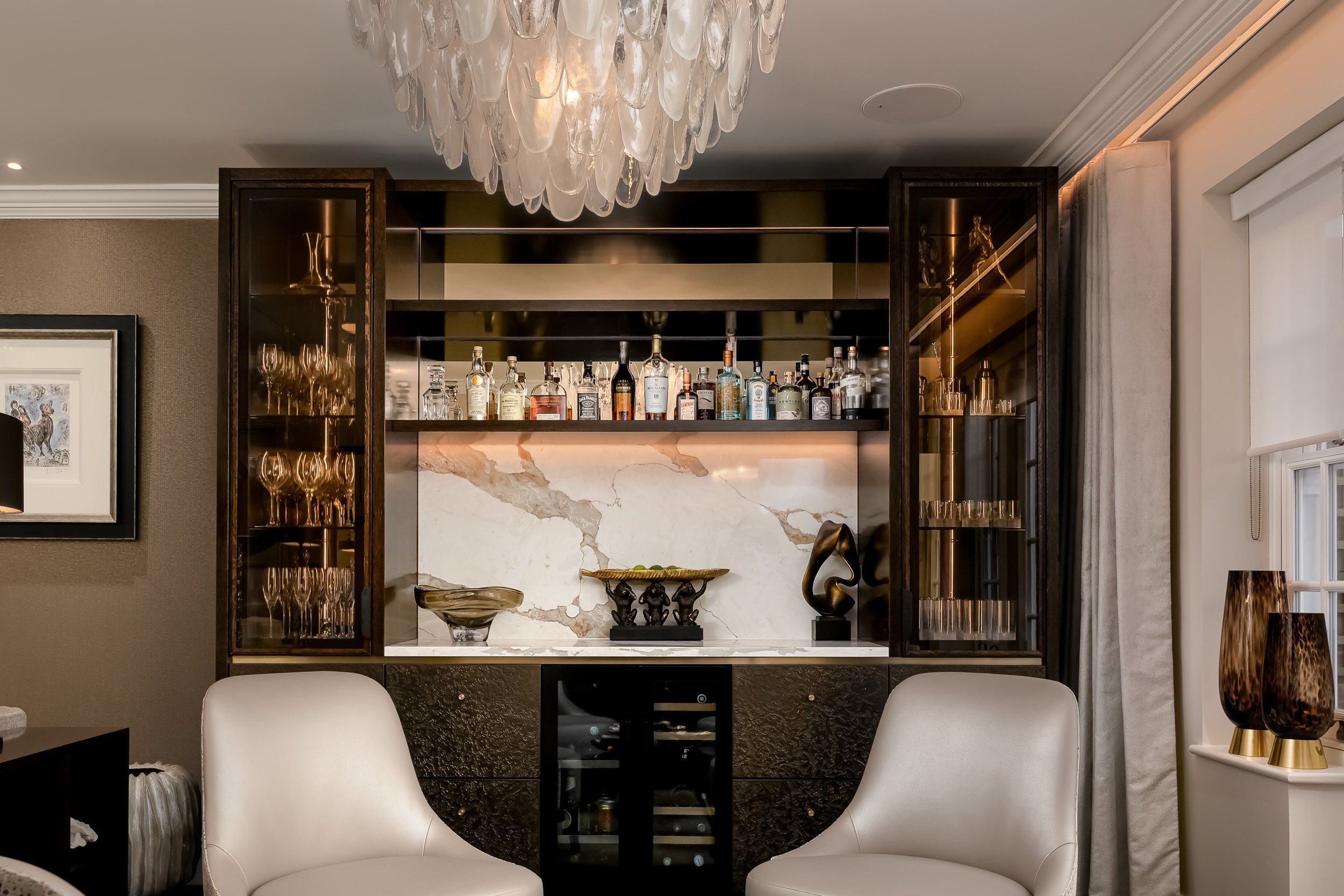Bespoke home bar with dark fitted cabinetry, illuminated shelving, stone detailing and decorative chandelier.